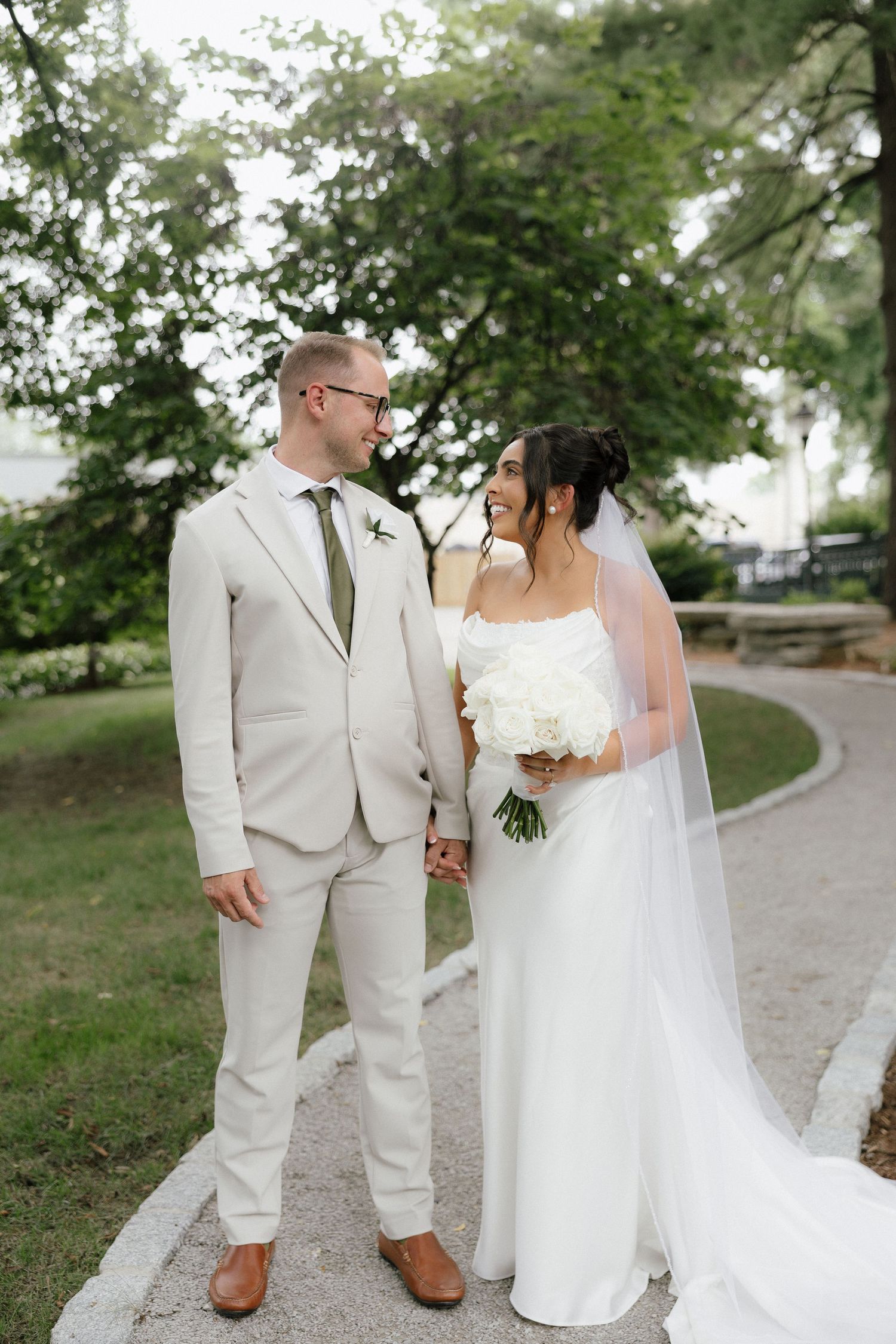 Bride and groom smile at each other on a garden path, she holds white flowers, he wears a tan suit with green tie.