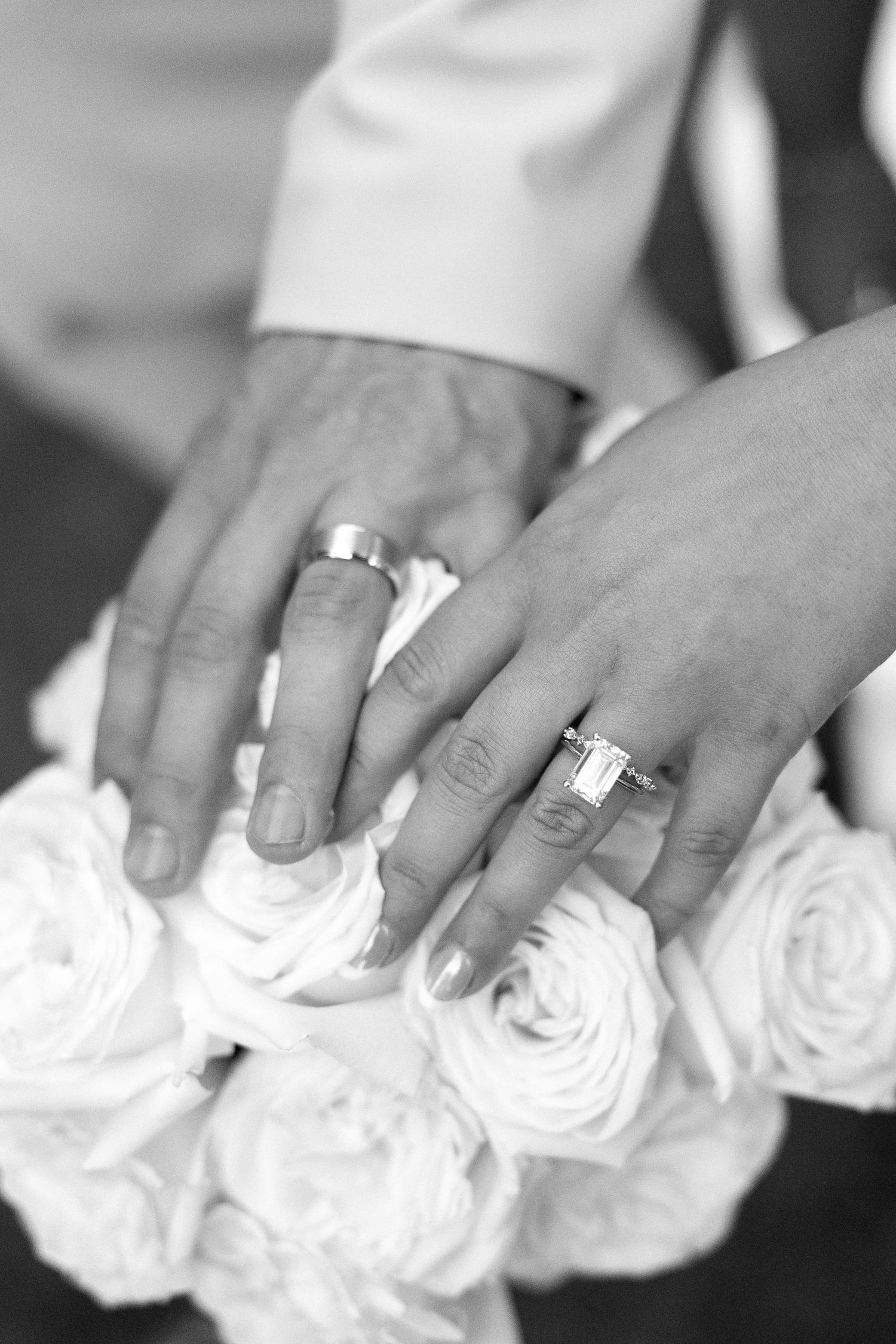 Black and white close-up of newlyweds hands with wedding rings resting on white rose bouquet.