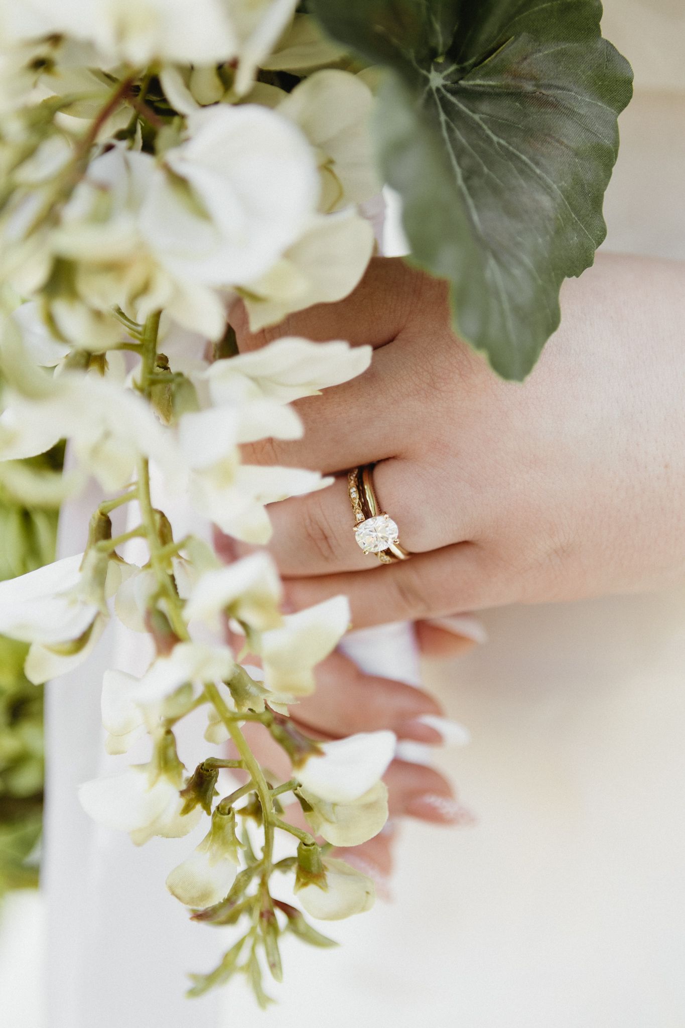 Close-up of an engagement ring on a hand holding delicate white flowers and green leaves.
