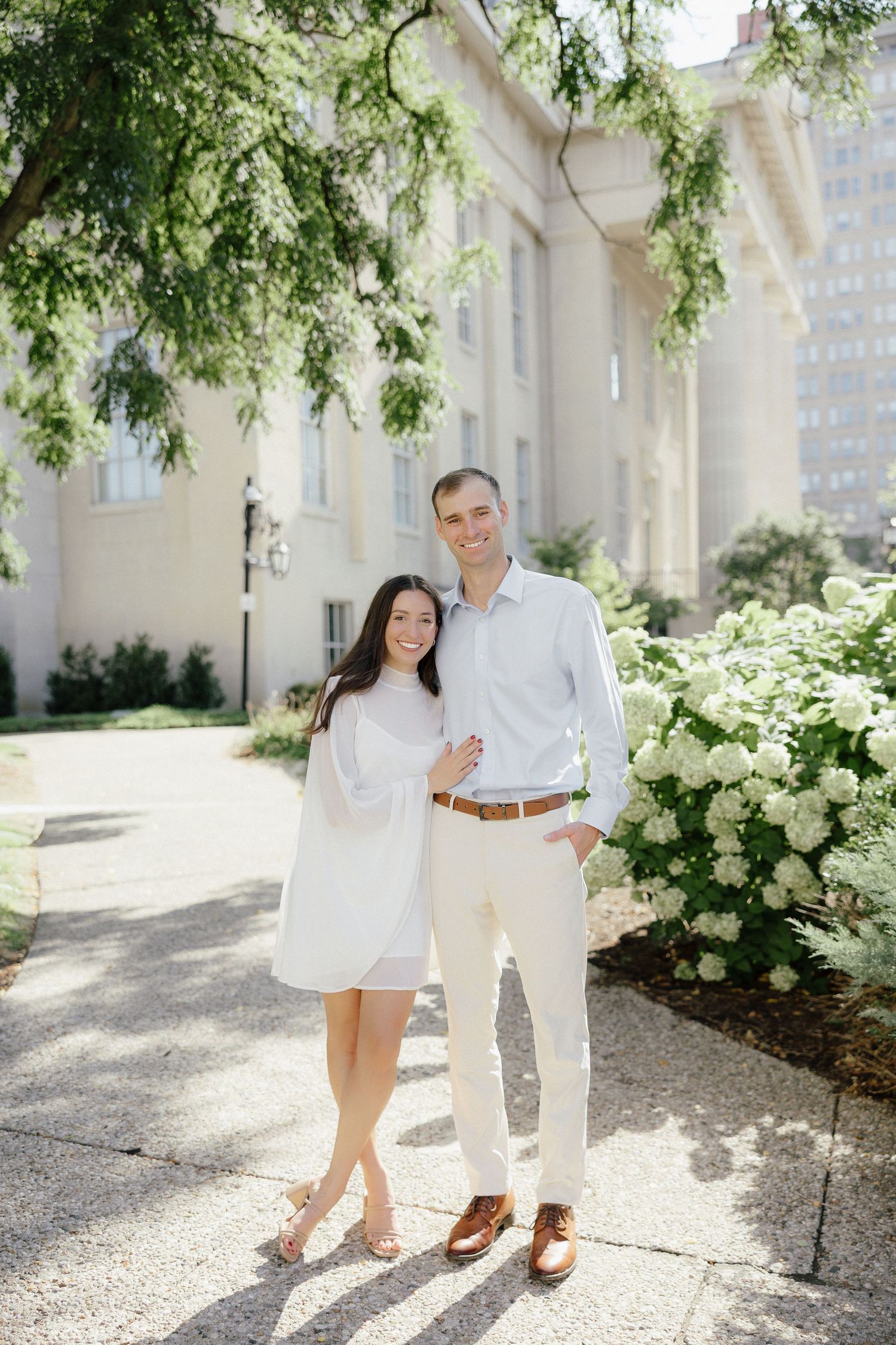 Couple posing together near blooming white hydrangeas outside a classic building, woman in white dress and man in light pants.