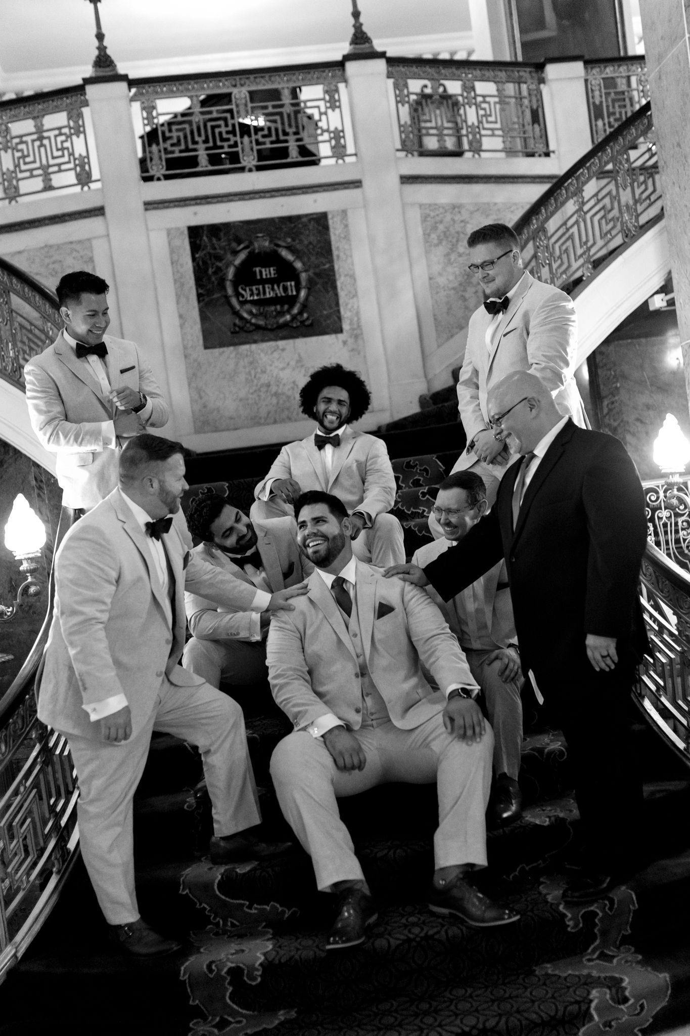 A group of men in light-colored suits sit and stand on an ornate staircase with decorative railings in a black and white photo.