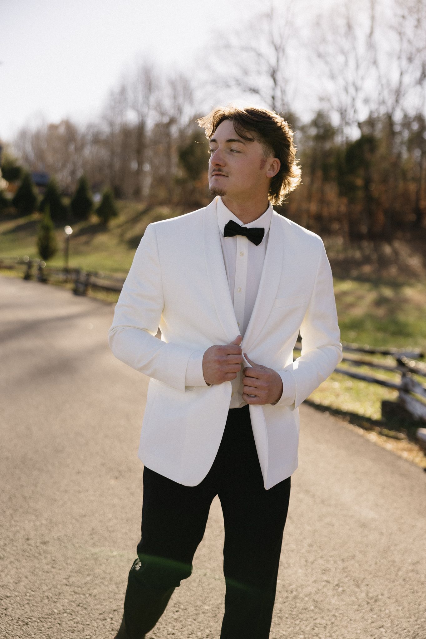 A groom in a white dinner jacket and bow tie poses on a sunlit path.
