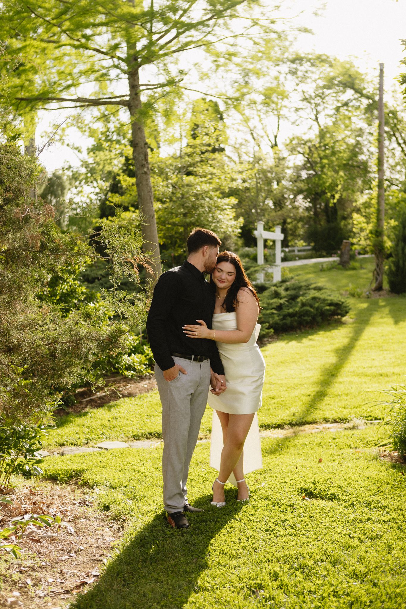 Couple shares romantic moment on grassy hillside path surrounded by lush green trees in springtime.