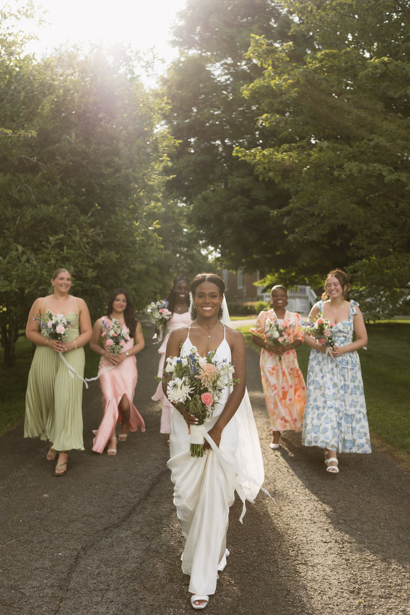 A joyful wedding party walks down a tree-lined path during a sun-drenched photo session.