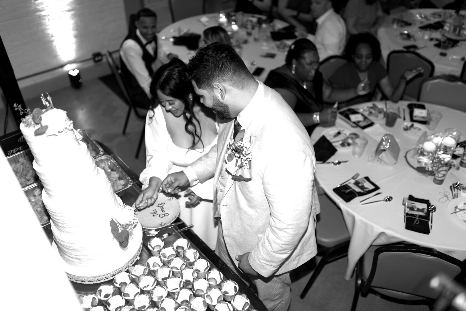 Black and white photograph of people gathered around a table at a celebration with cupcakes being served.