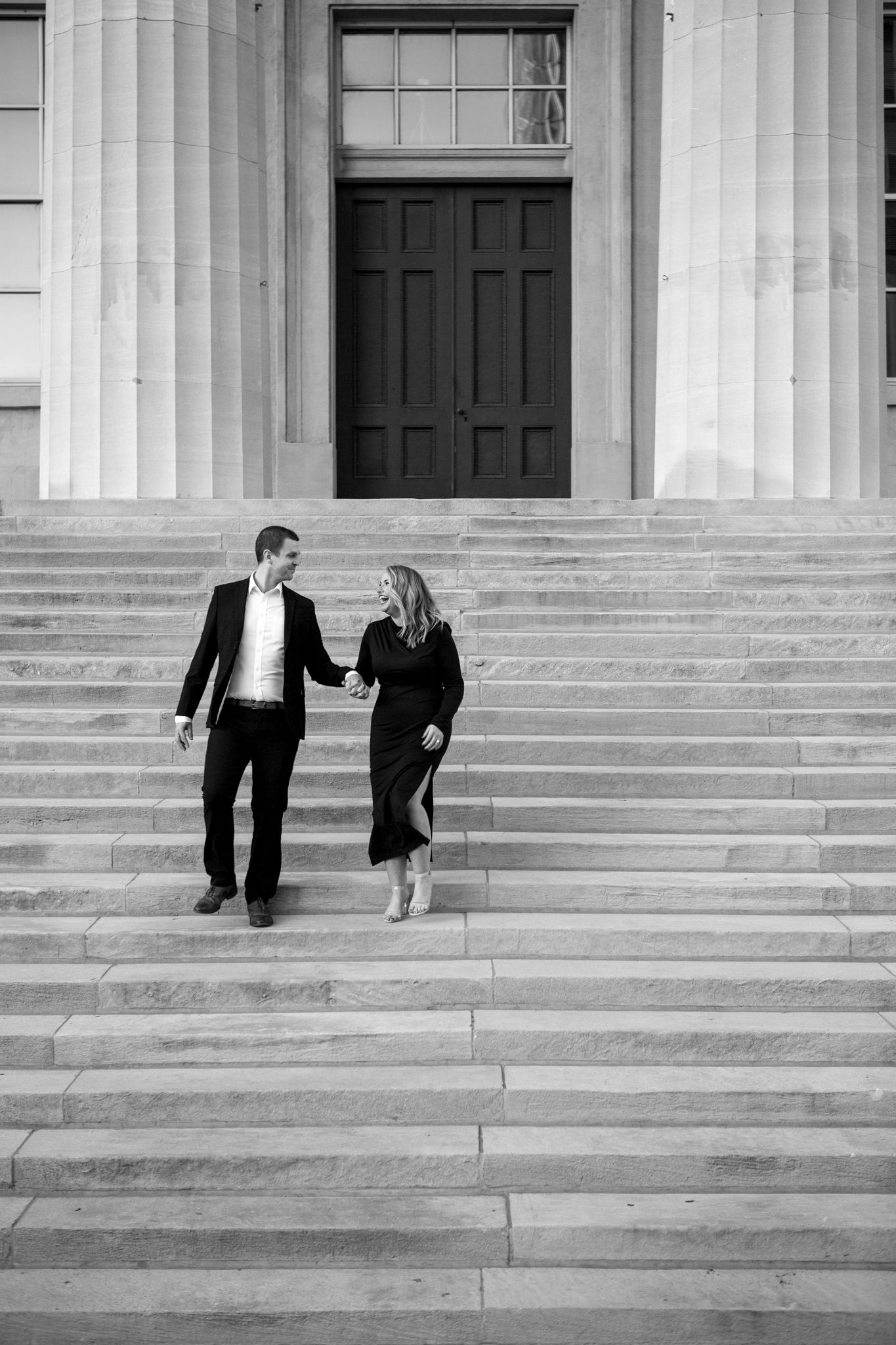 A couple walks up grand stone steps between classical columns in an elegant black and white photograph.
