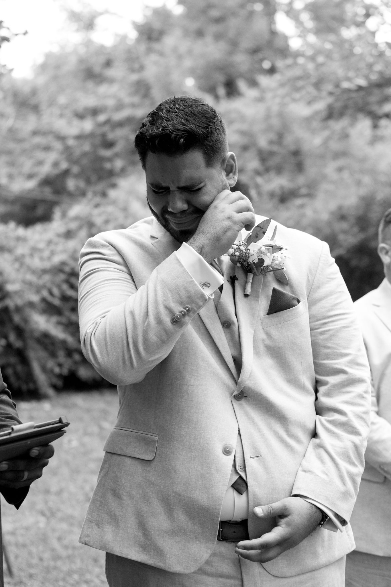 Black and white emotional wedding moment showing groom in light suit wiping tears during ceremony outdoors.