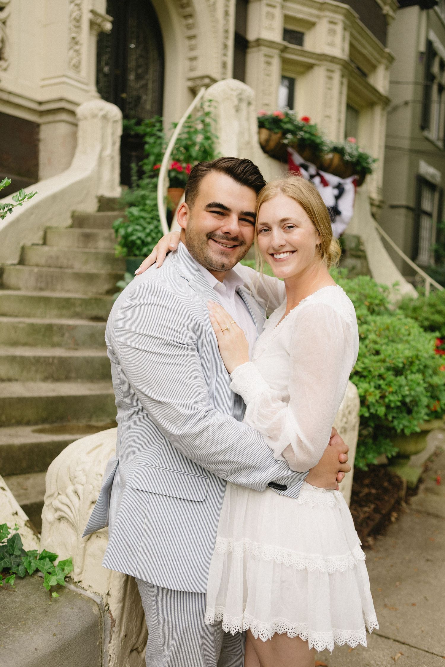 Smiling couple embracing outdoors in wedding attire, man in gray suit and woman in white lace dress, in front of elegant building.