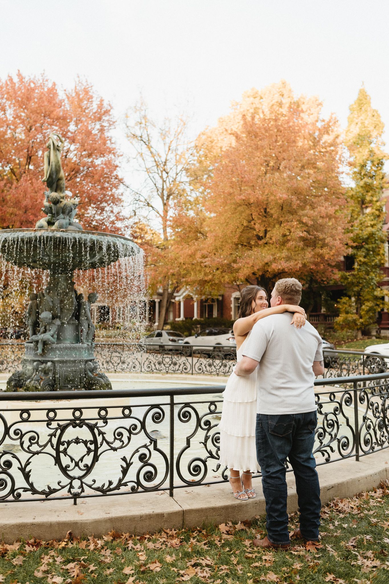 Couple embraces by decorative iron railing with autumn foliage and gazebo in background.
