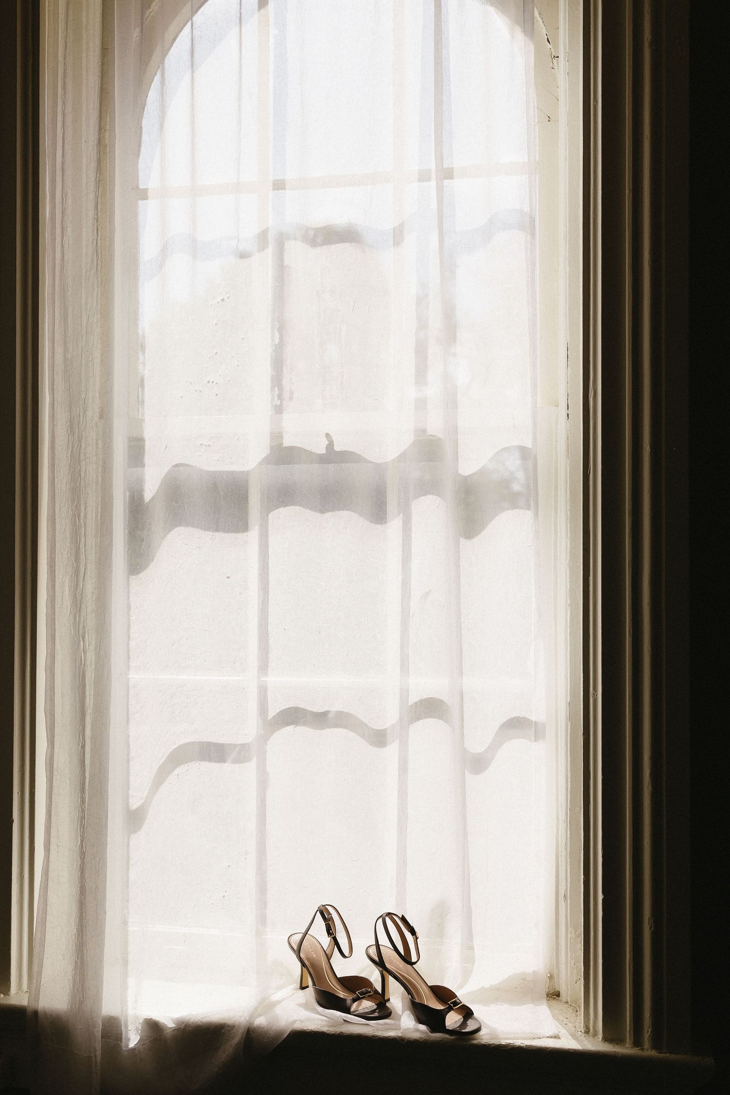 Elegant wedding shoes silhouetted against sheer curtains in an arched window.