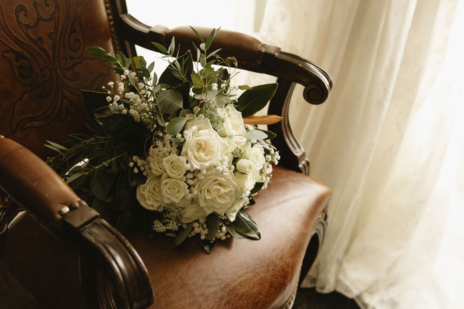 A white floral bouquet sits elegantly on a brown leather chair against a light-colored curtain backdrop.