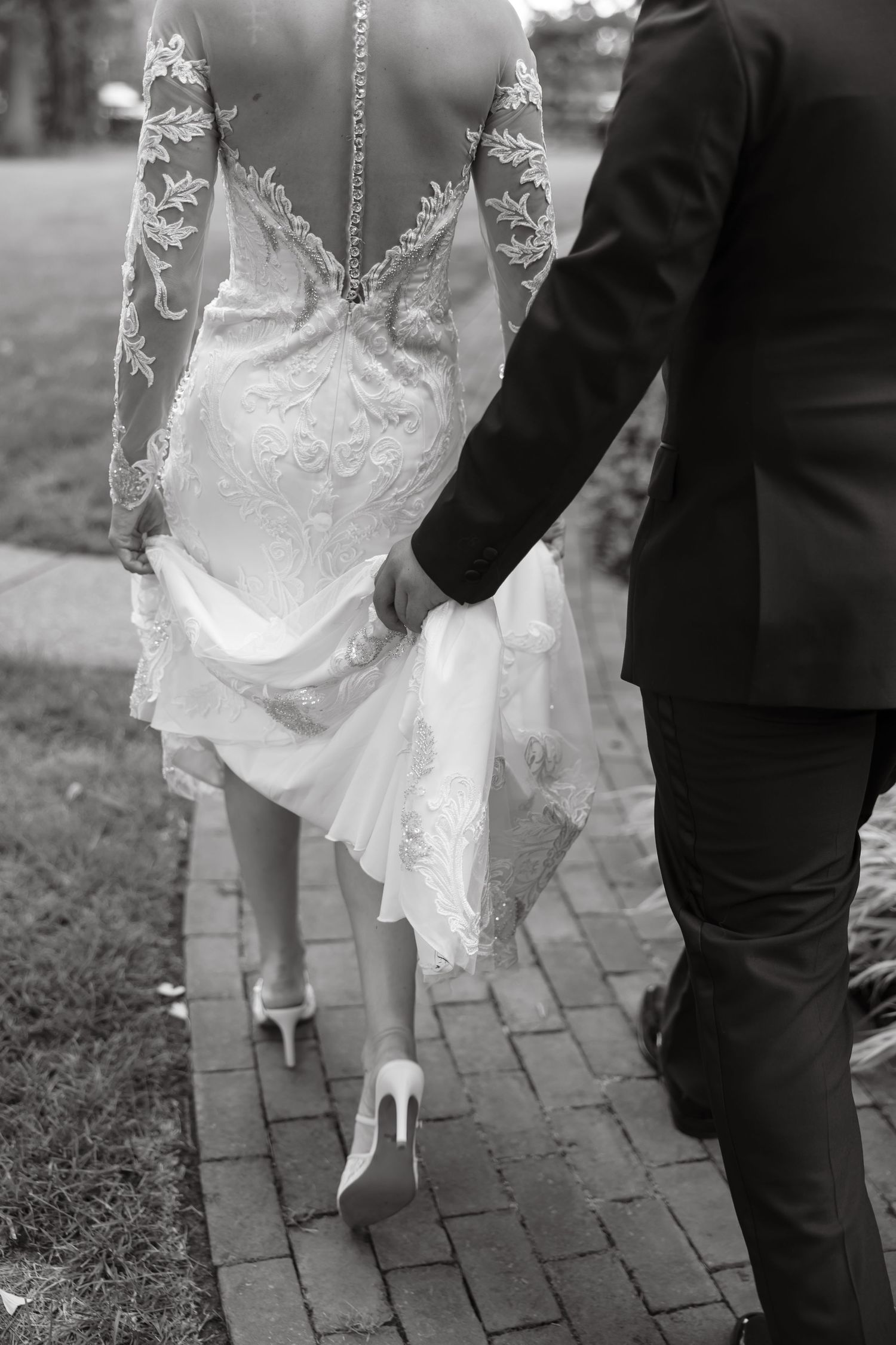Black and white photo of bride in lace long-sleeve gown walking brick path with groom holding her dress train.