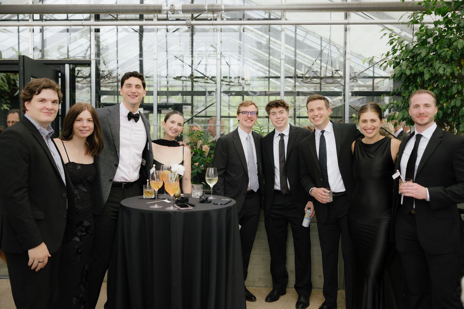 Group of wedding guests in formal black attire posing together in a greenhouse venue.