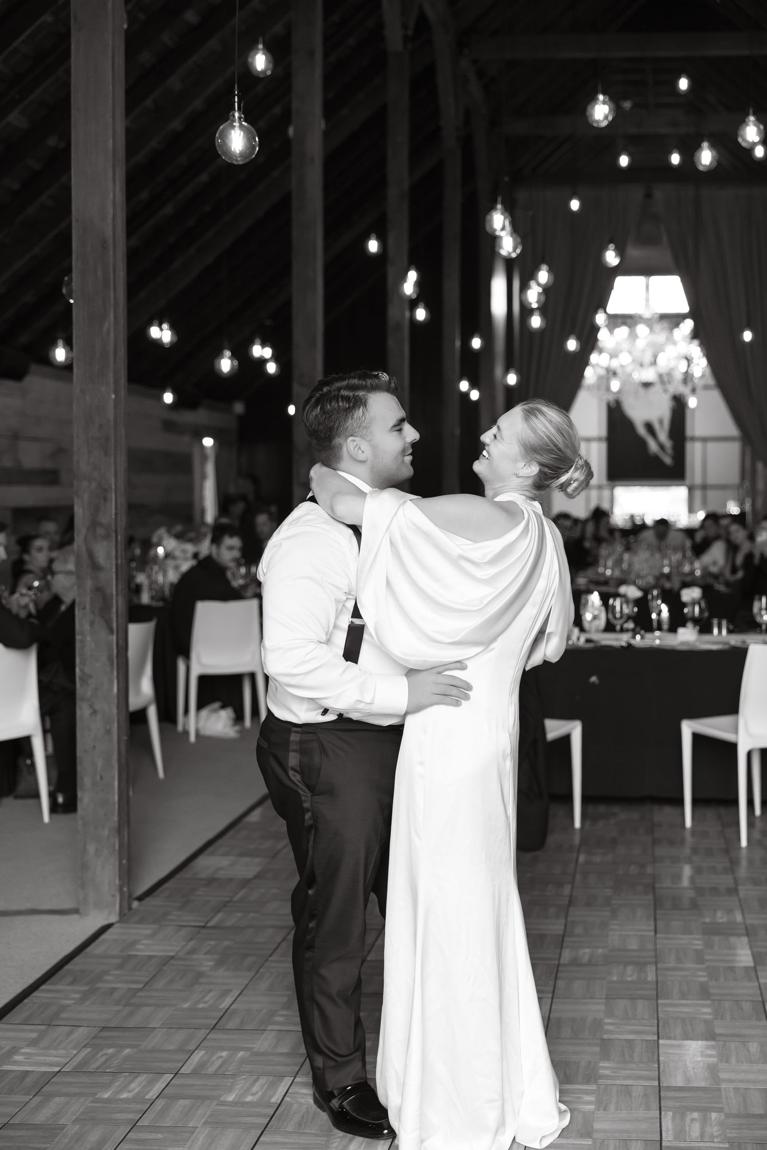 Bride and groom share romantic first dance in black and white photo inside rustic barn with globe string lights.