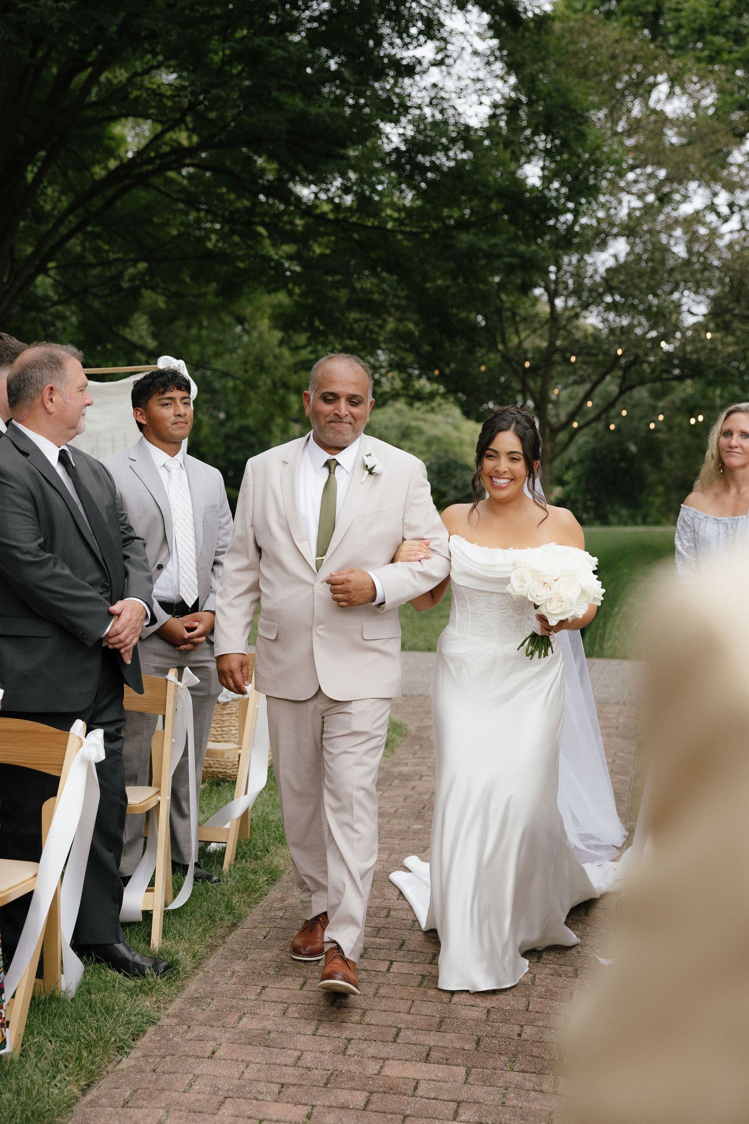 Bride walks down outdoor aisle with her father, wearing a white gown and holding white bouquet at garden wedding.