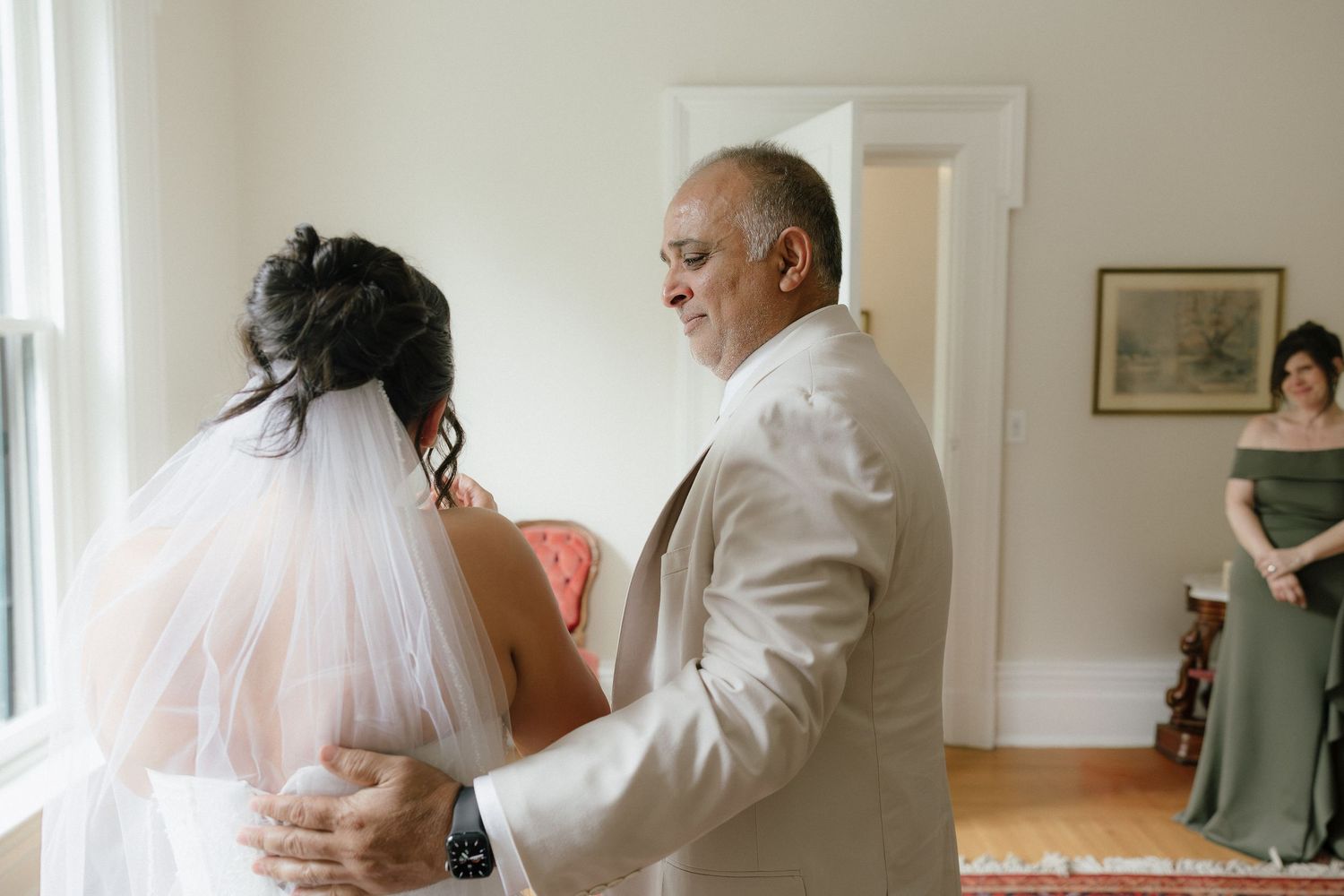 Father and bride share an emotional first look moment, with a bridesmaid in sage green dress visible in background.