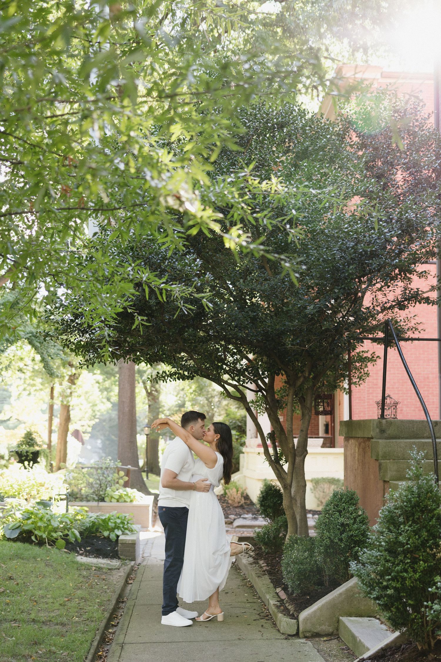 Couple embracing on a garden path surrounded by lush green trees and shrubs near a brick building.