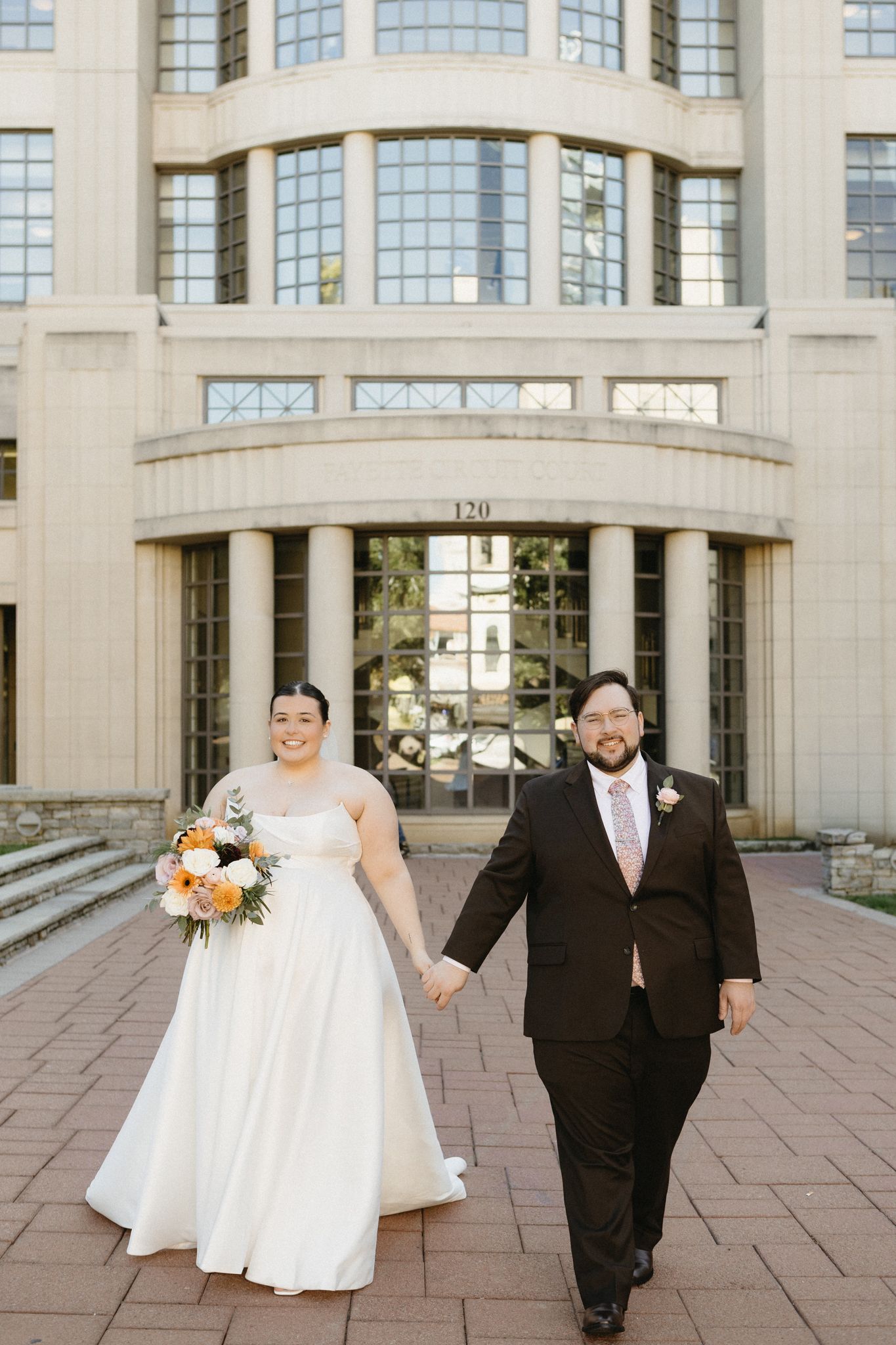 A wedding couple poses together on a stone courtyard in front of a grand building with large windows.