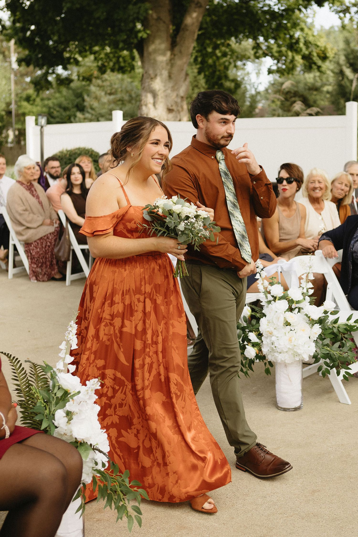 Wedding party in burnt orange dresses and olive suits dancing at outdoor celebration.