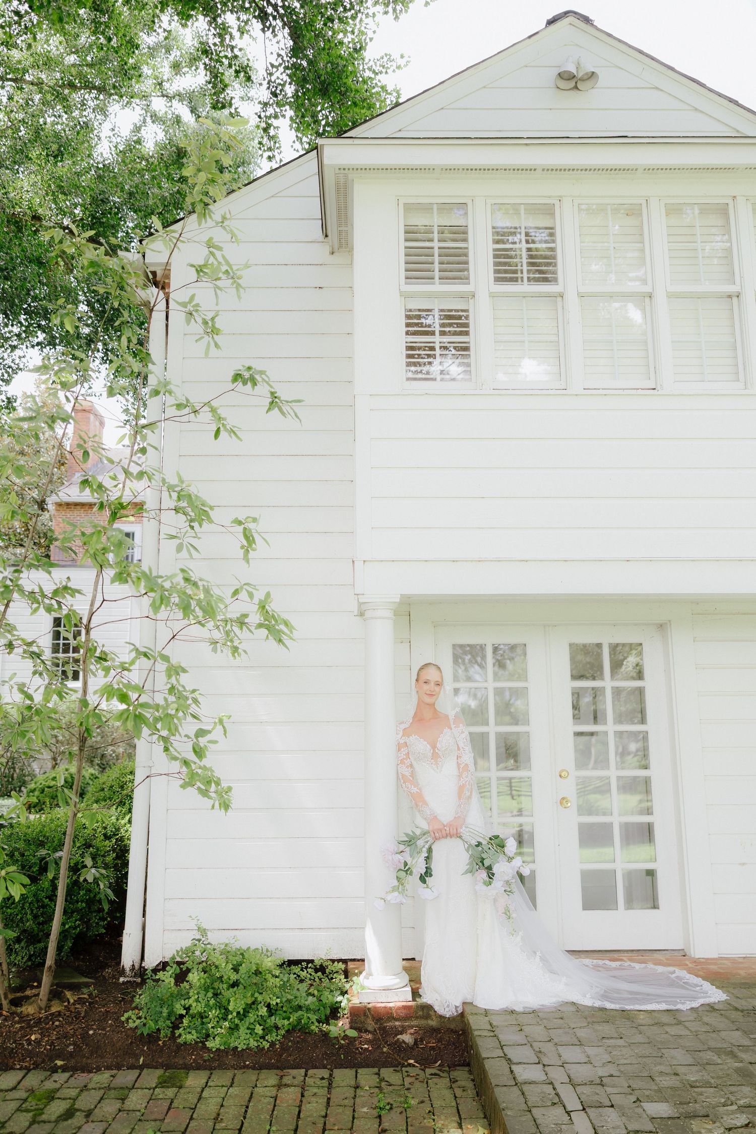 Bride in lace wedding gown holding bouquet stands before charming white two-story colonial home surrounded by lush greenery.