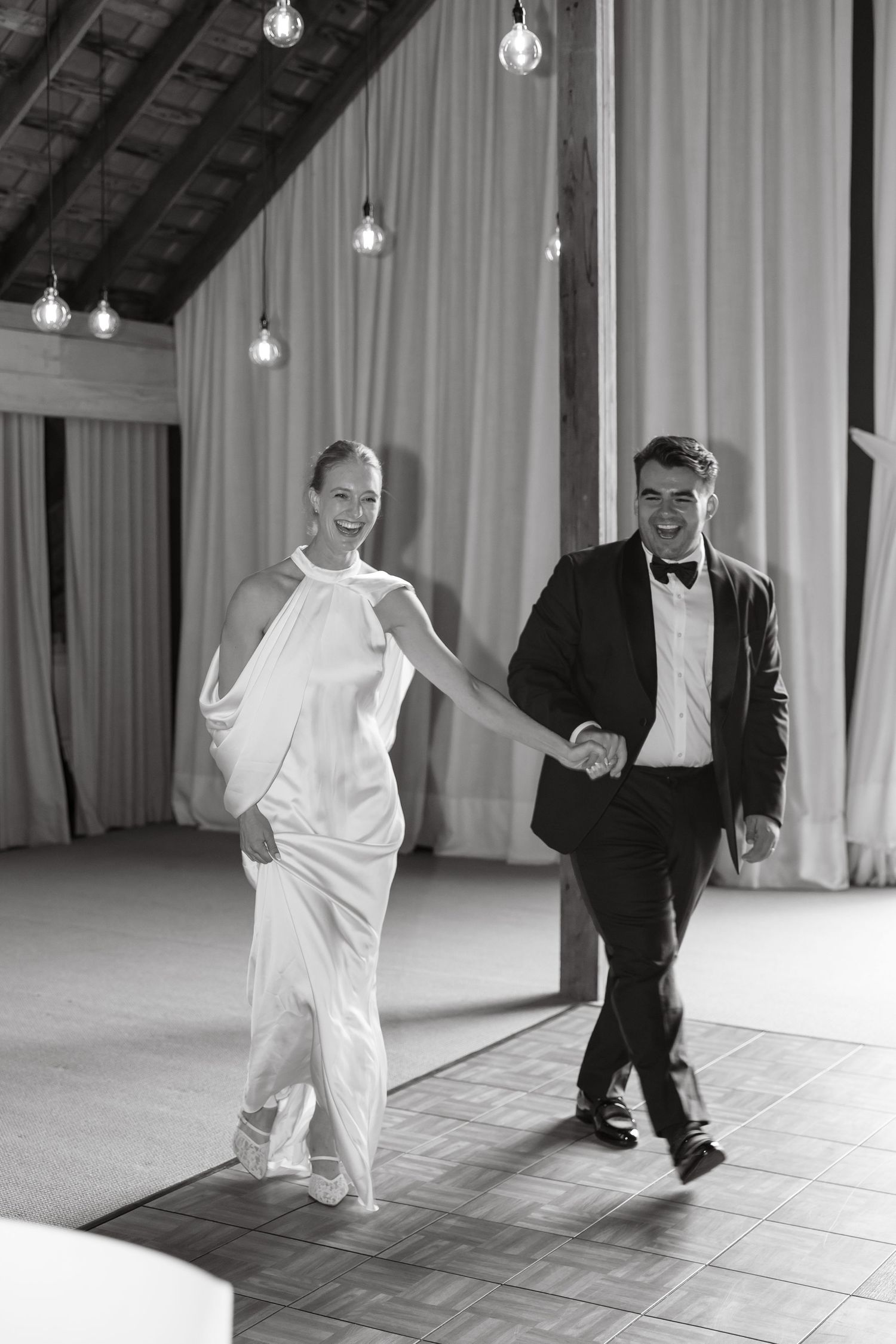 Black and white photo of smiling bride and groom making grand entrance at wedding reception venue.