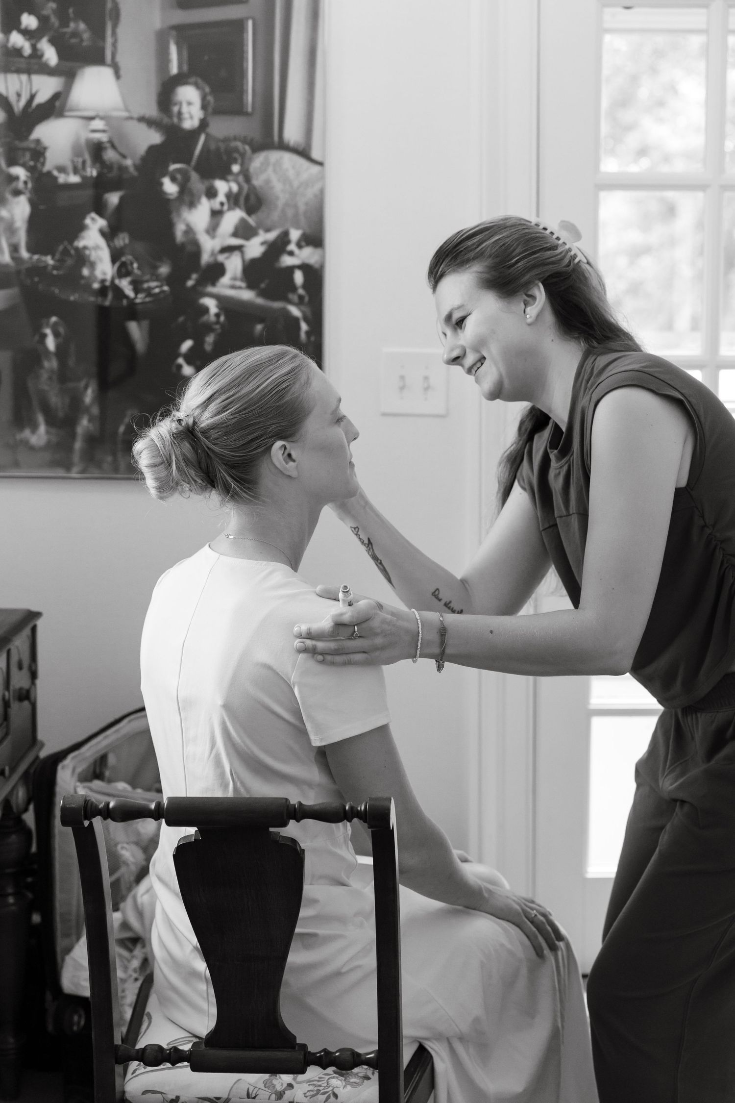 A makeup artist applies makeup to a bride seated in a chair during wedding preparations in black and white.