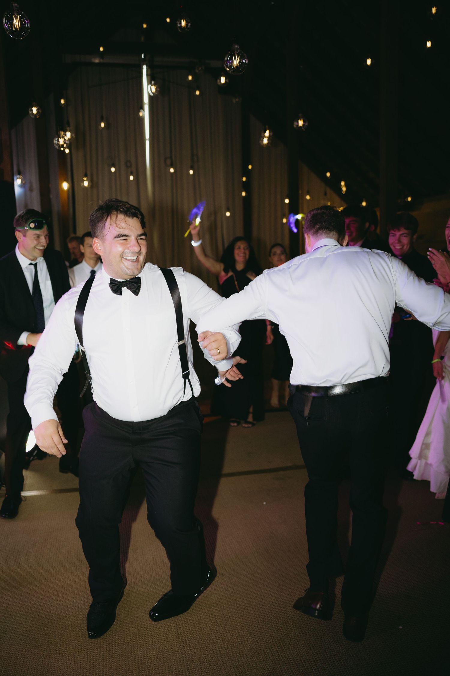 Energetic wedding guest in suspenders dancing with groom under Edison bulbs at lively barn reception party.