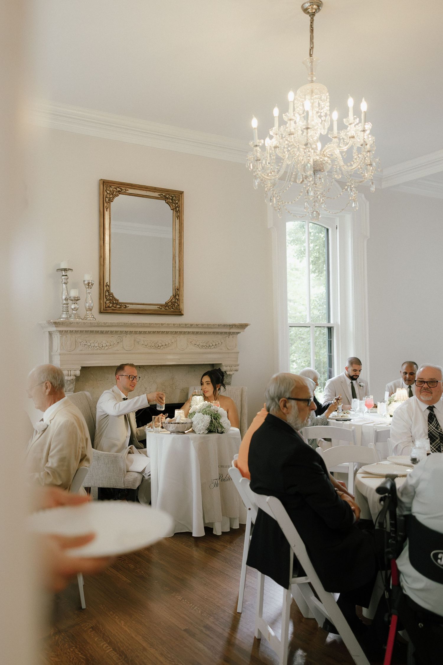 Wedding reception dinner in an elegant white room with crystal chandelier, fireplace, and gold mirror.