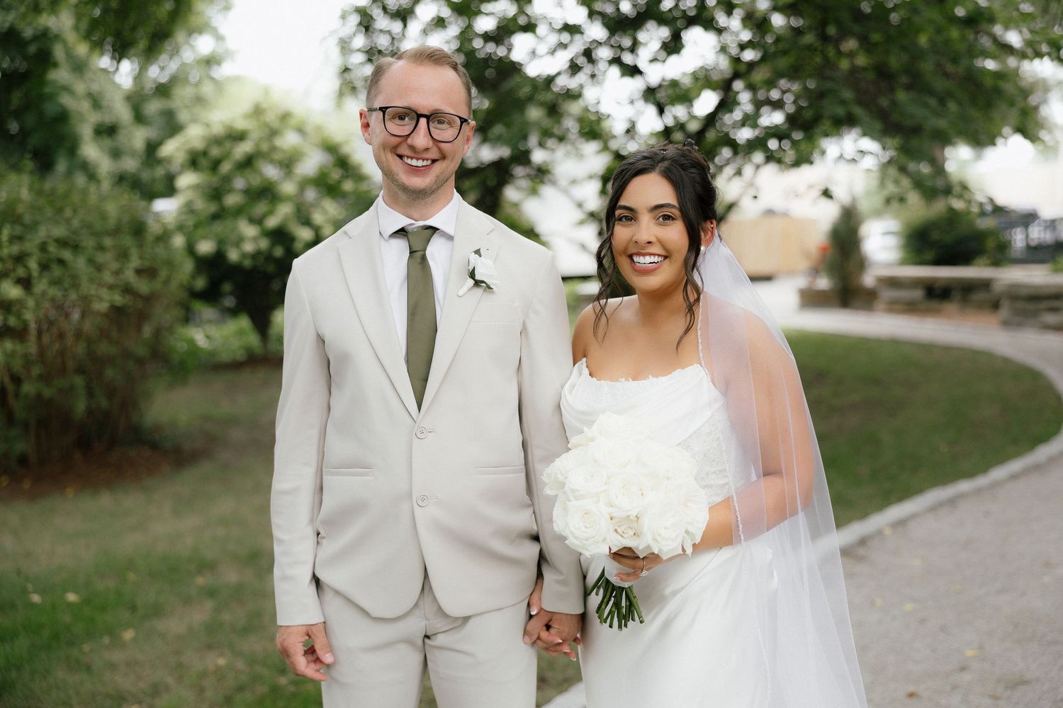Smiling bride and groom holding hands outdoors, bride in white gown with veil and bouquet, groom in beige suit.