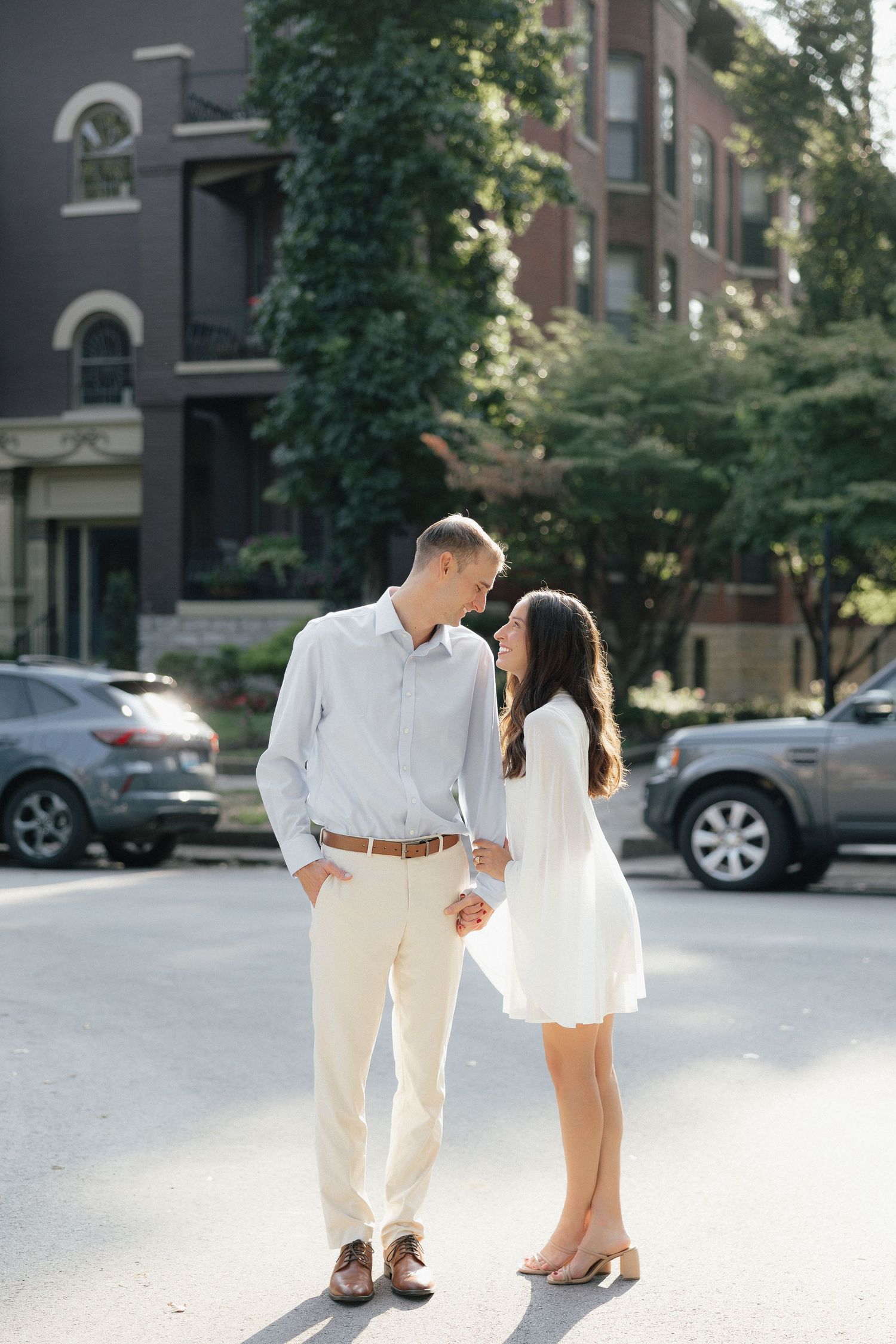 Couple holding hands and gazing at each other on a tree-lined urban street in coordinating white outfits.