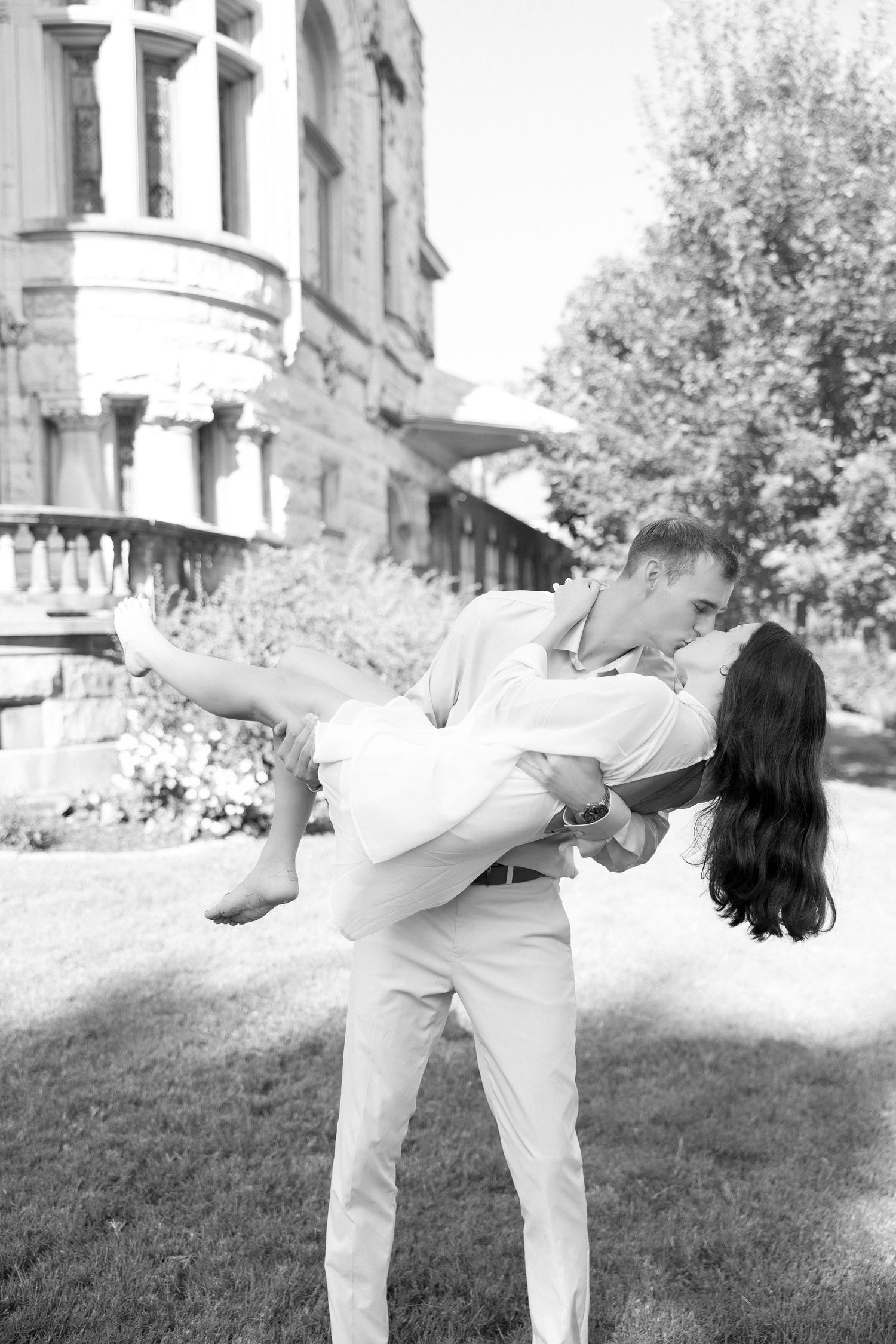 A romantic couple shares a kiss as the man lifts the woman in a black and white outdoor photo.