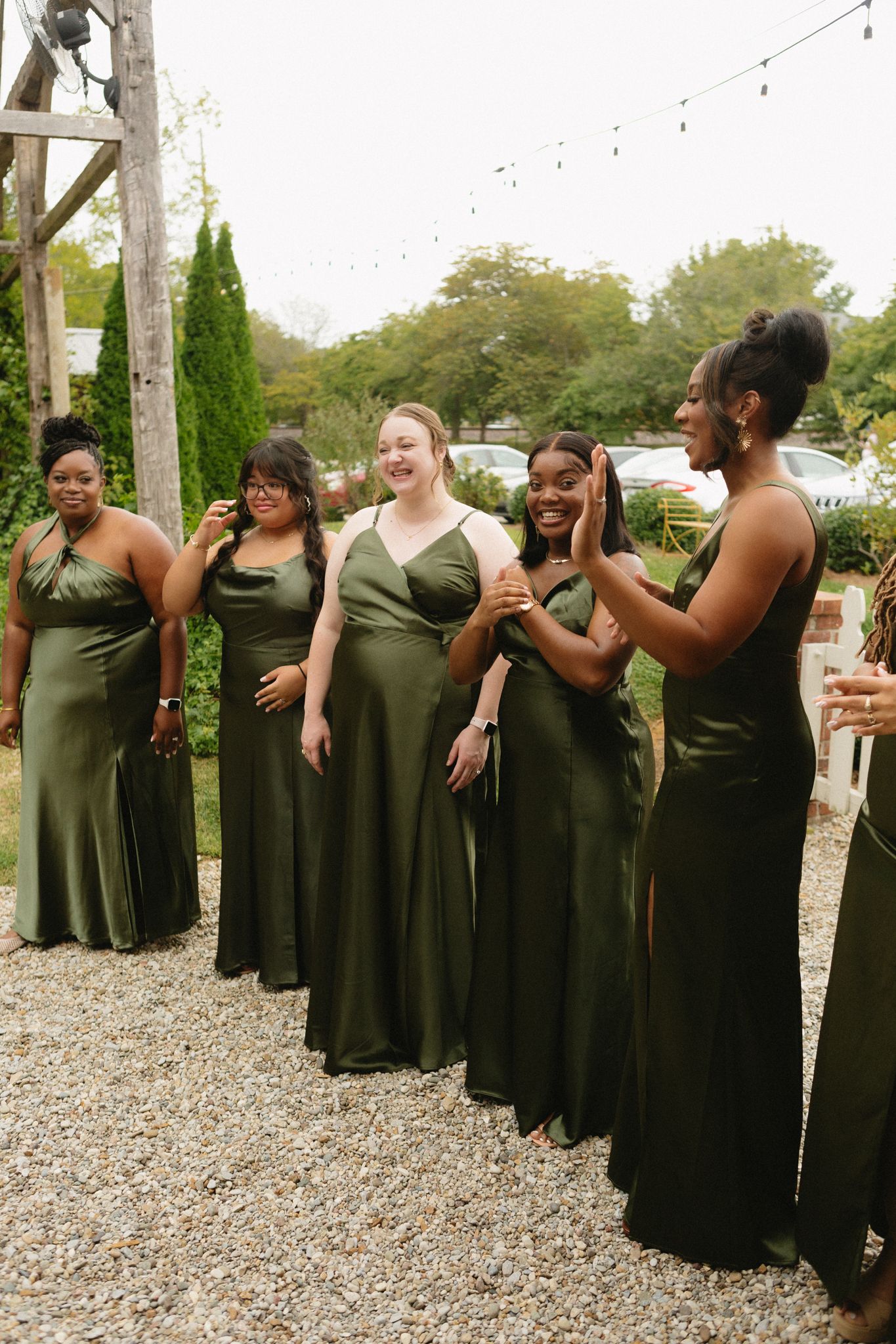 Bridesmaids in elegant olive green floor-length dresses share a candid laughing moment on a gravel path outdoors.