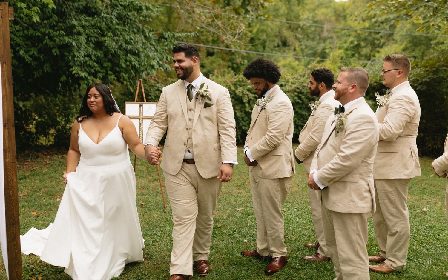 A bride and groom with their wedding party members wearing beige suits stand together on grass during an outdoor ceremony.
