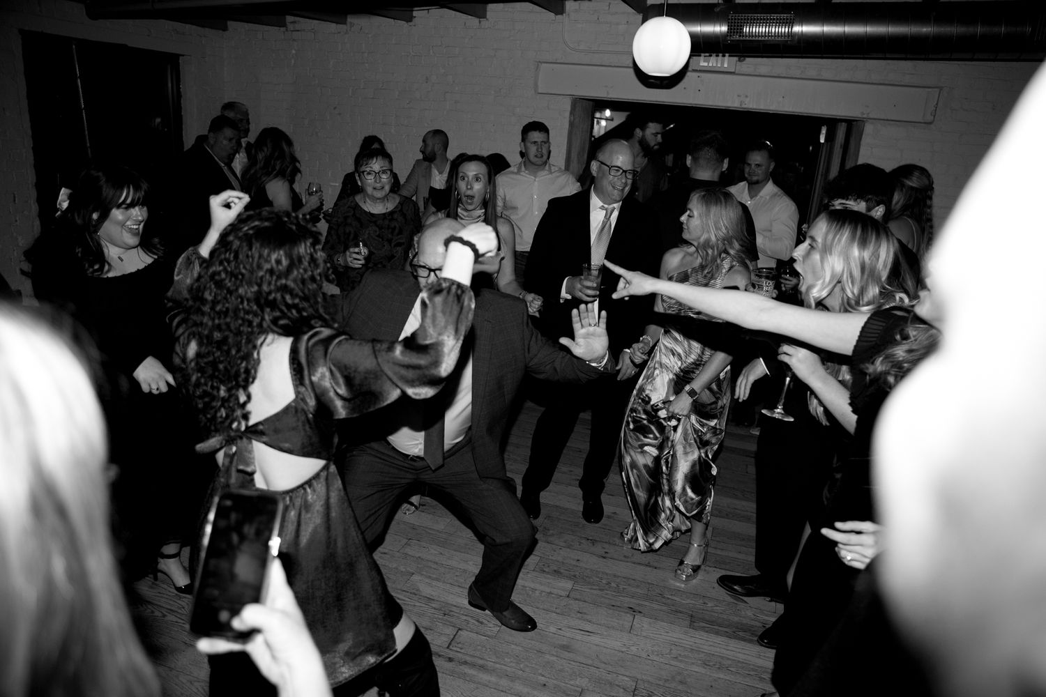 Wedding guests dancing enthusiastically at a reception on a wooden dance floor in black and white.