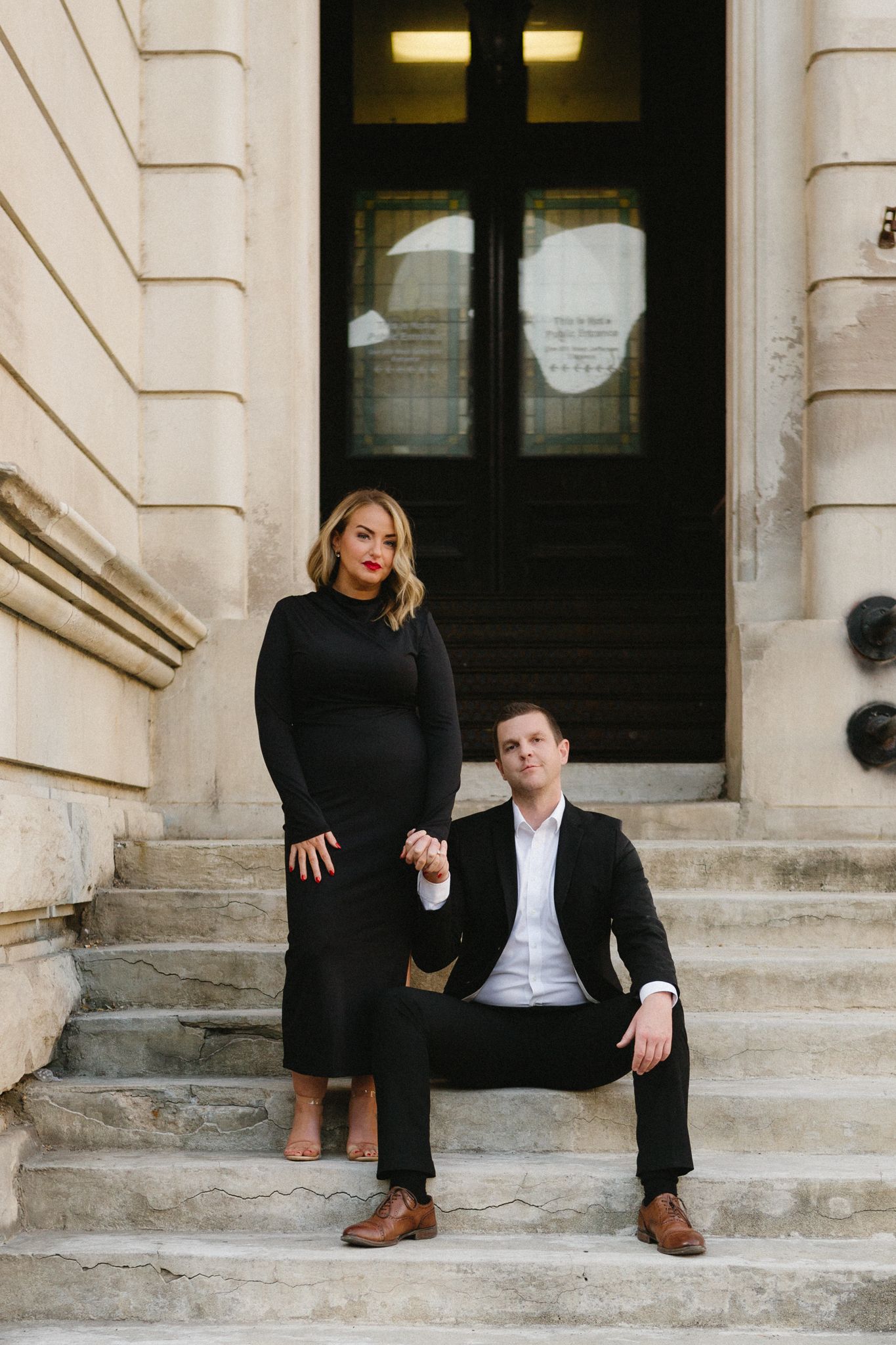 A couple in formal black attire poses on stone steps in front of a vintage doorway.