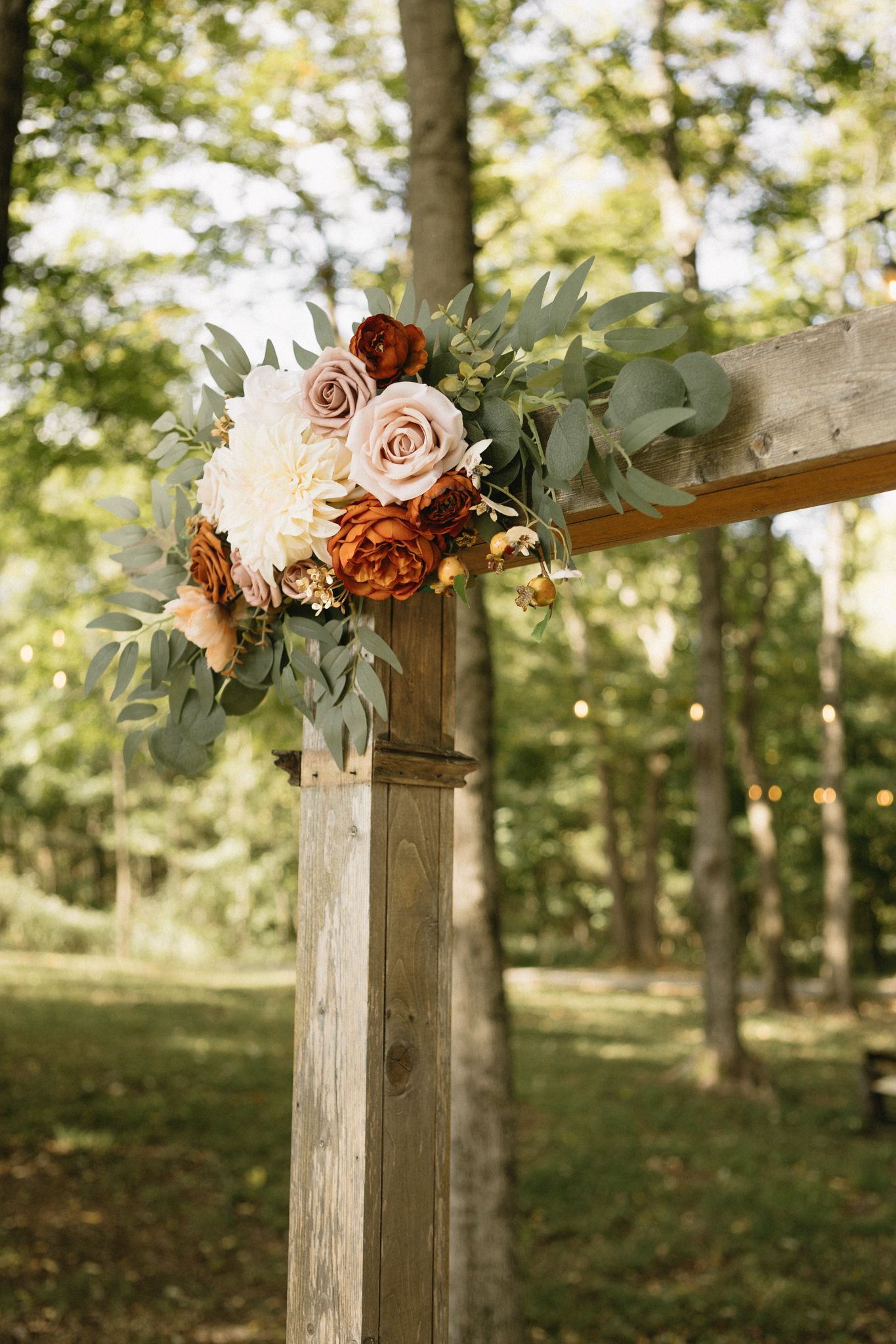 Rustic wooden wedding arch decorated with blush and burgundy flowers and greenery in an outdoor woodland setting.