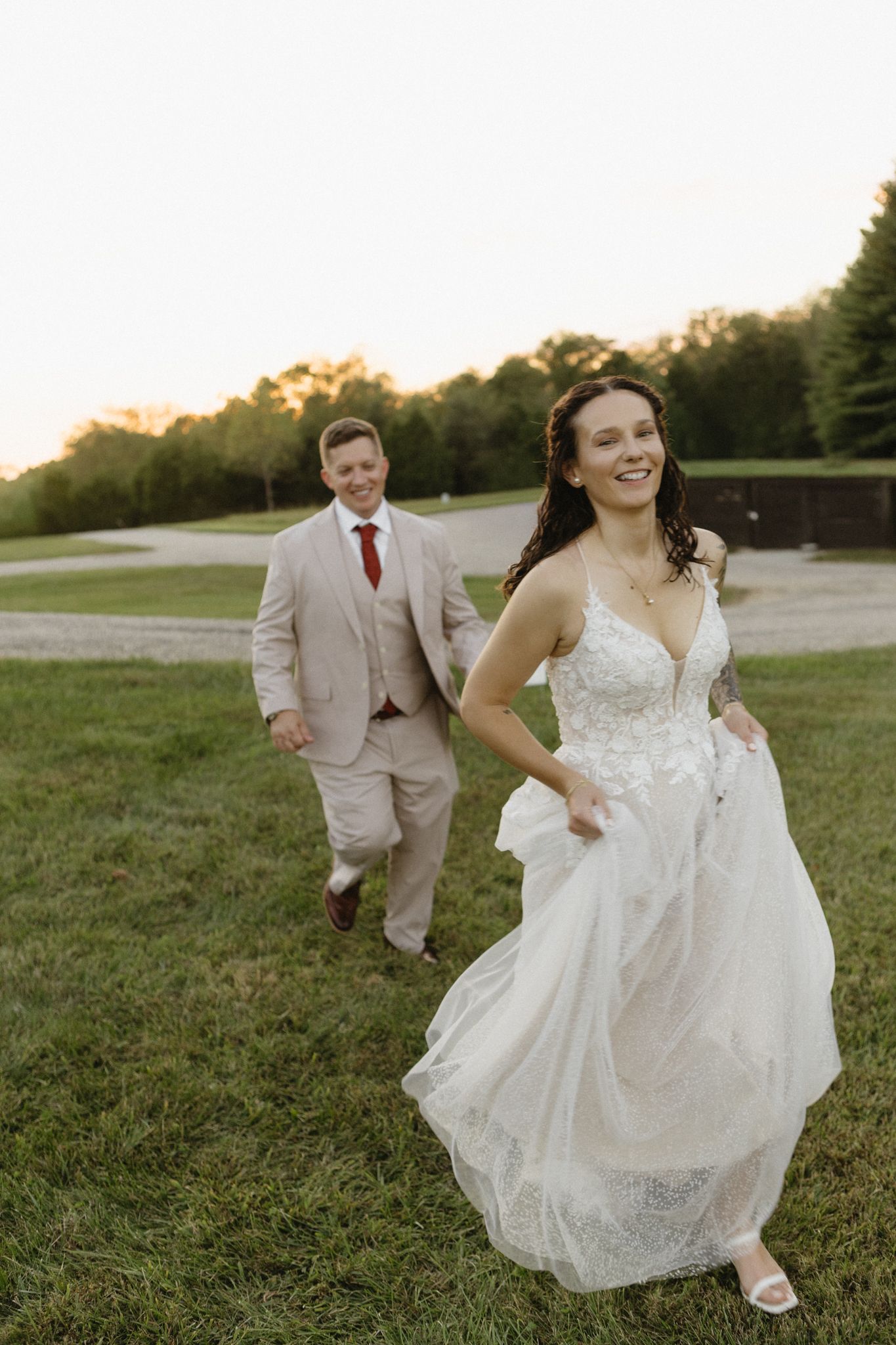 Newlyweds playfully run through a grassy field at sunset, bride in lace gown and groom in tan suit.