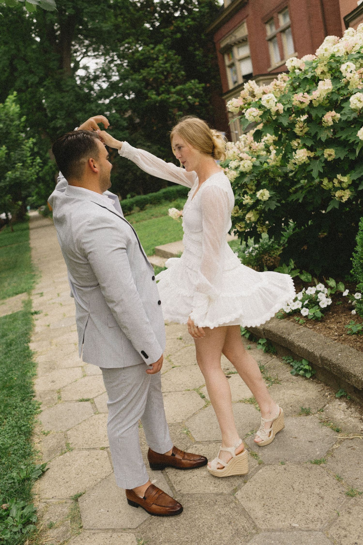 Couple dancing outdoors near blooming white flowers, woman in white dress and man in gray suit on stone path.