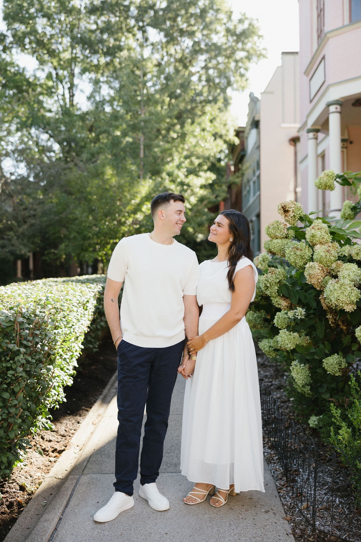 A couple holds hands and smiles at each other on a sunny sidewalk, surrounded by lush greenery and blooming hydrangeas.