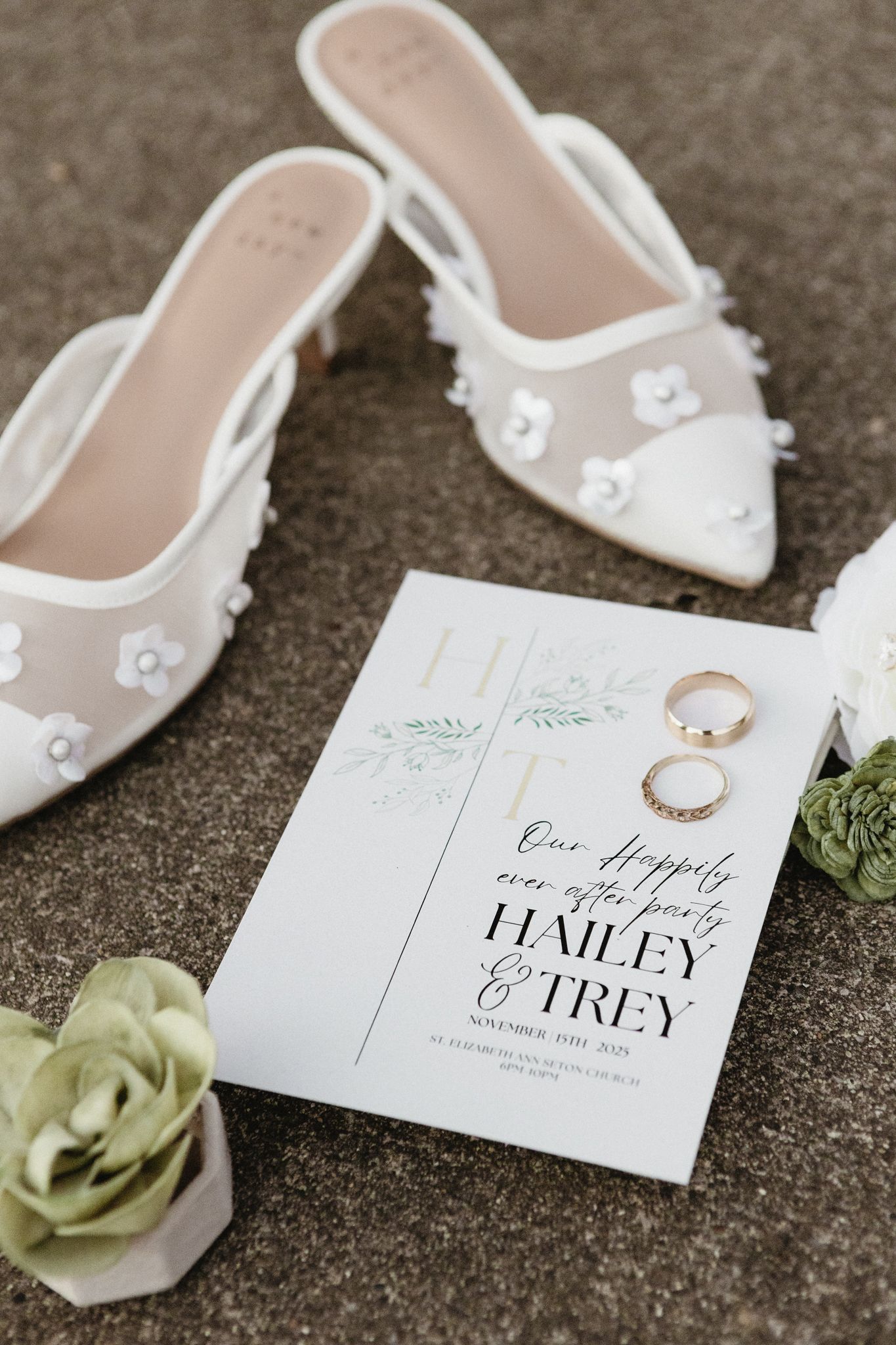 Wedding invitation and elegant white bridal shoes with rings displayed on a gray surface next to delicate green florals.