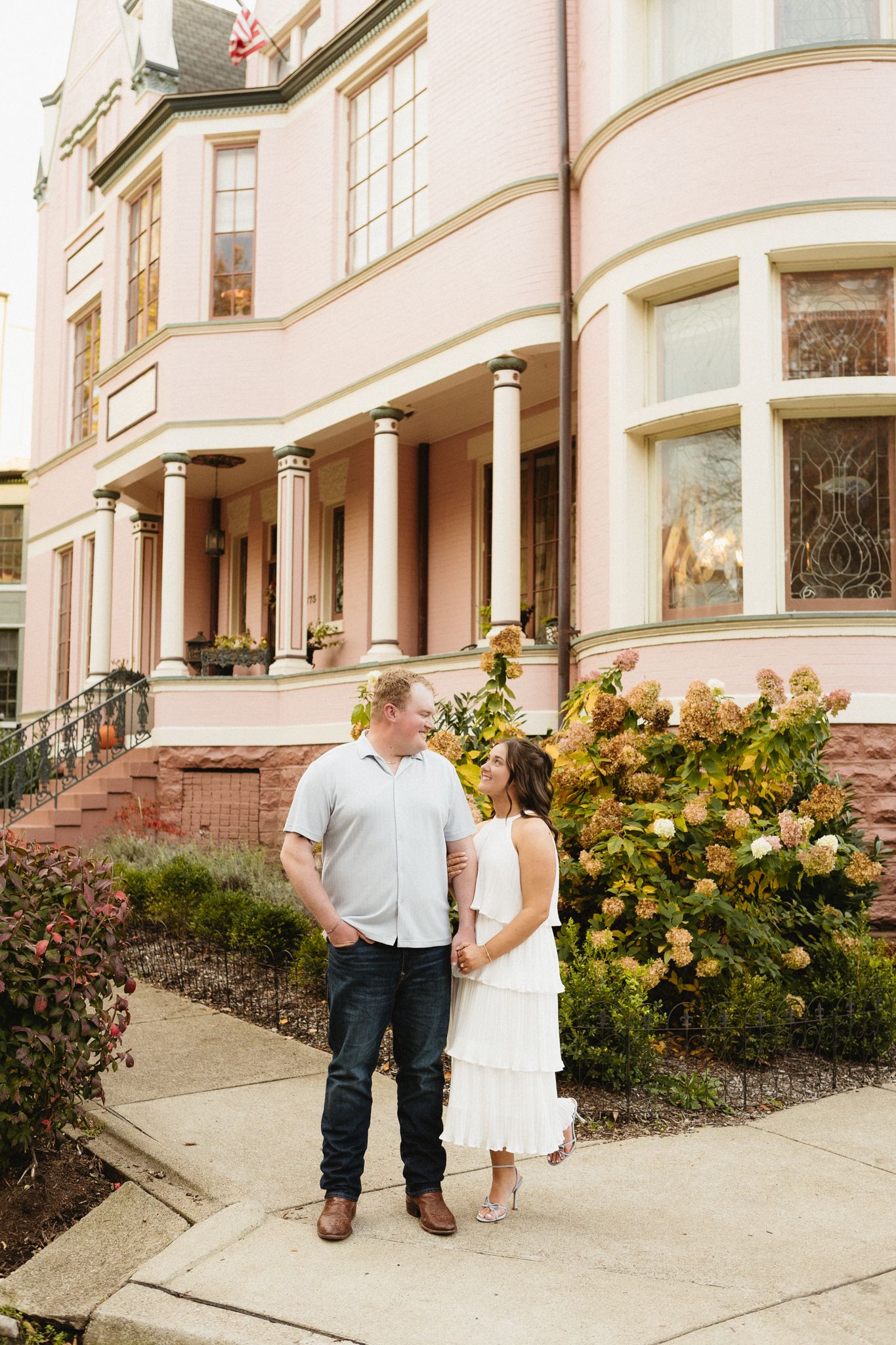 Romantic couple poses in front of elegant building with yellow roses blooming in landscaped garden.
