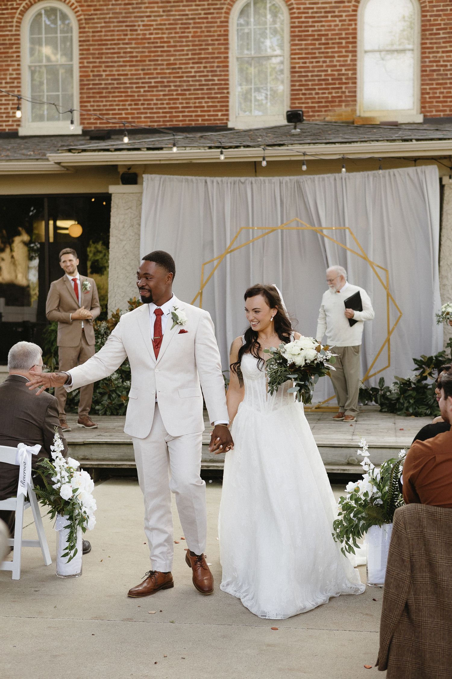 Newlyweds celebrate with guests in front of white drapes and geometric arch at outdoor venue.