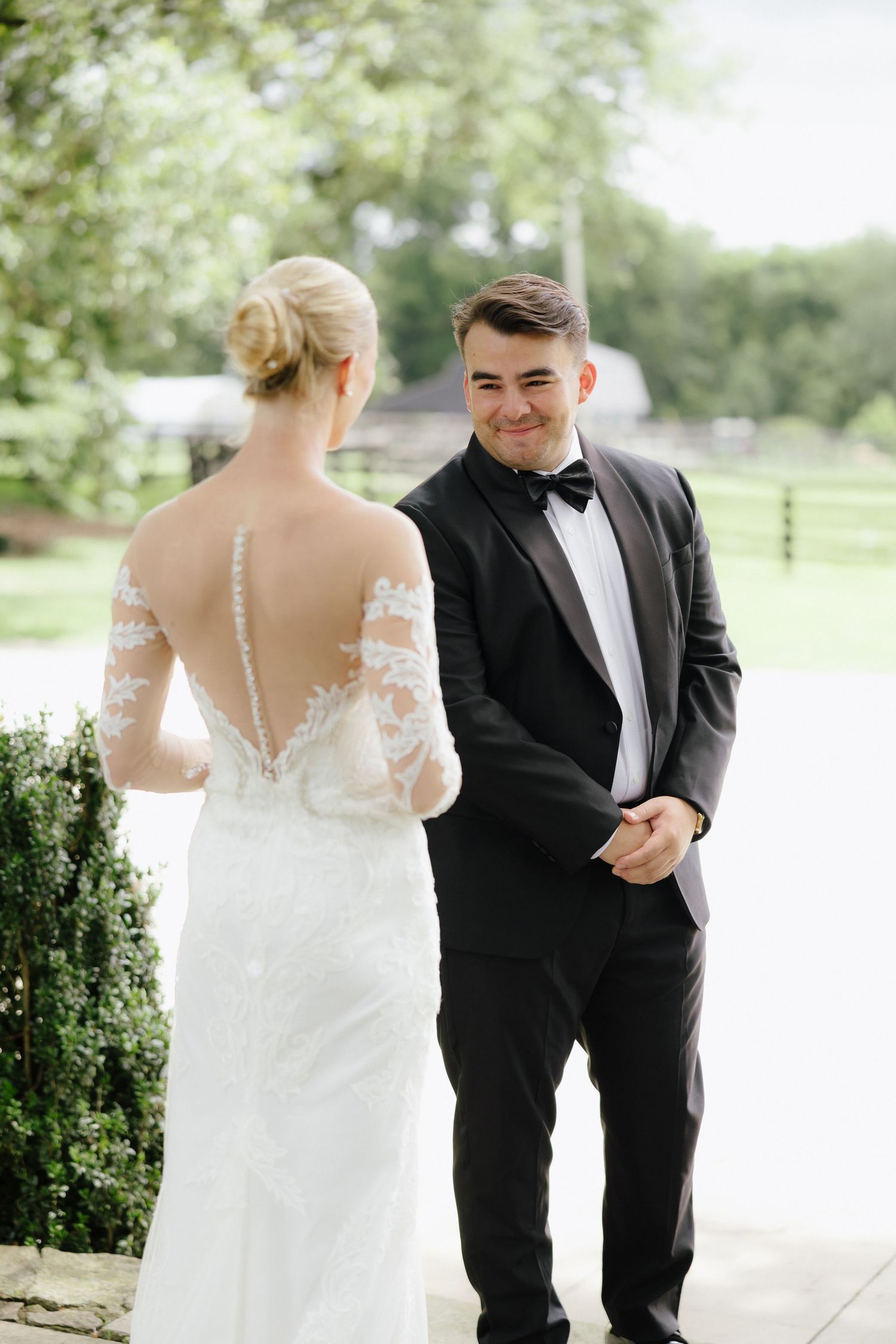 Groom smiling at bride during first look, bride in lace backless wedding gown, groom in black tuxedo.