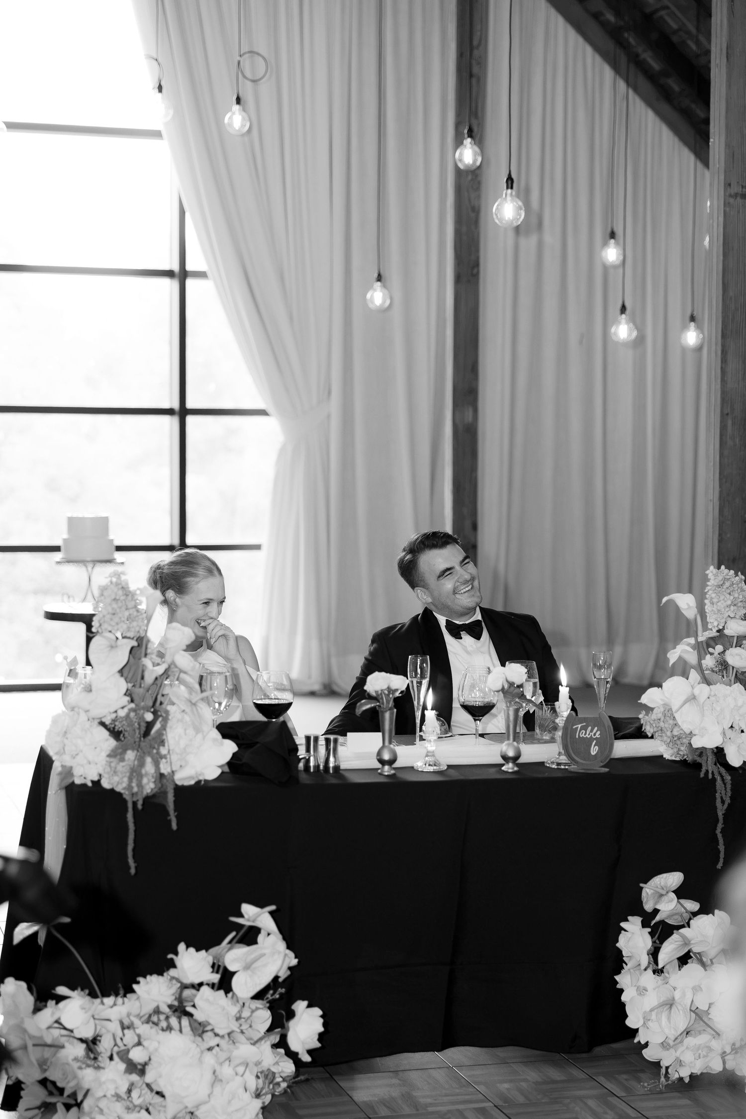 Black and white photo of bride and groom laughing at sweetheart table with floral arrangements.