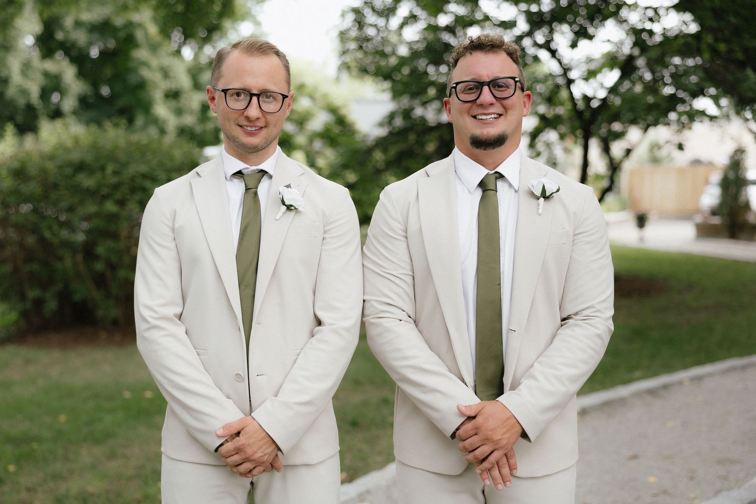 Two groomsmen in matching cream suits and olive green ties smile outdoors at a wedding.