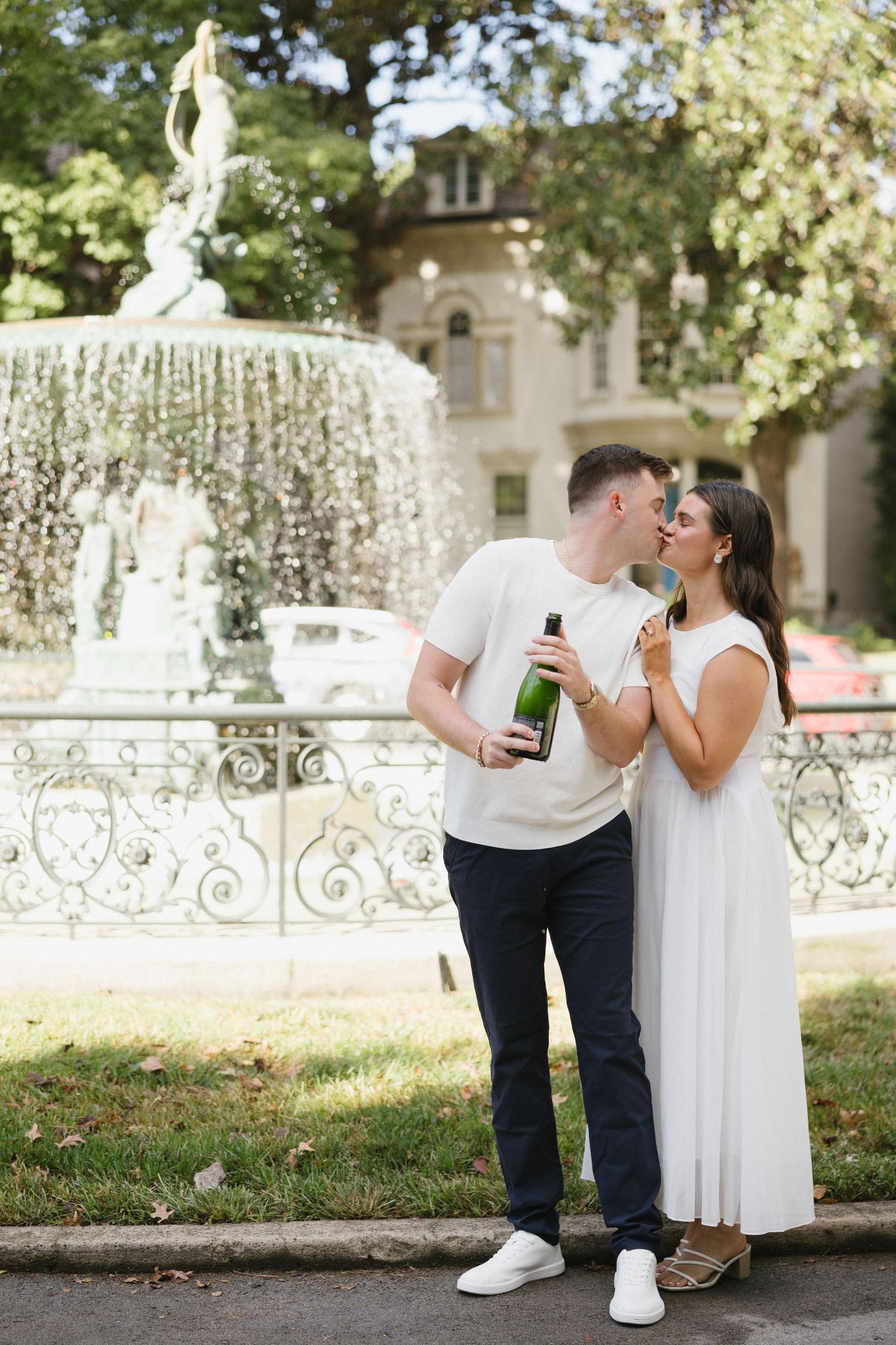 Romantic couple kissing by an ornate fountain, holding a champagne bottle, dressed in white outfits.