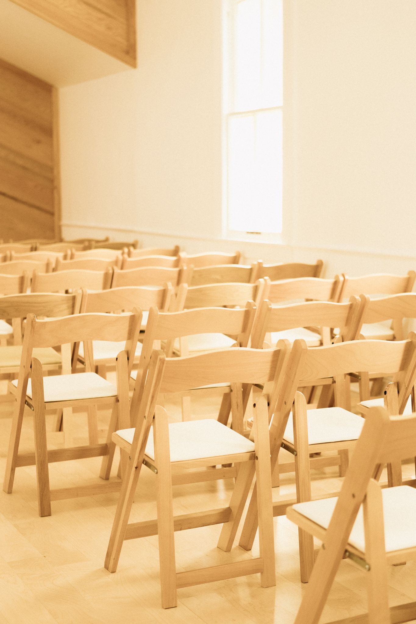 Rows of light wooden folding chairs with white cushions arranged in a bright room with natural lighting from windows.