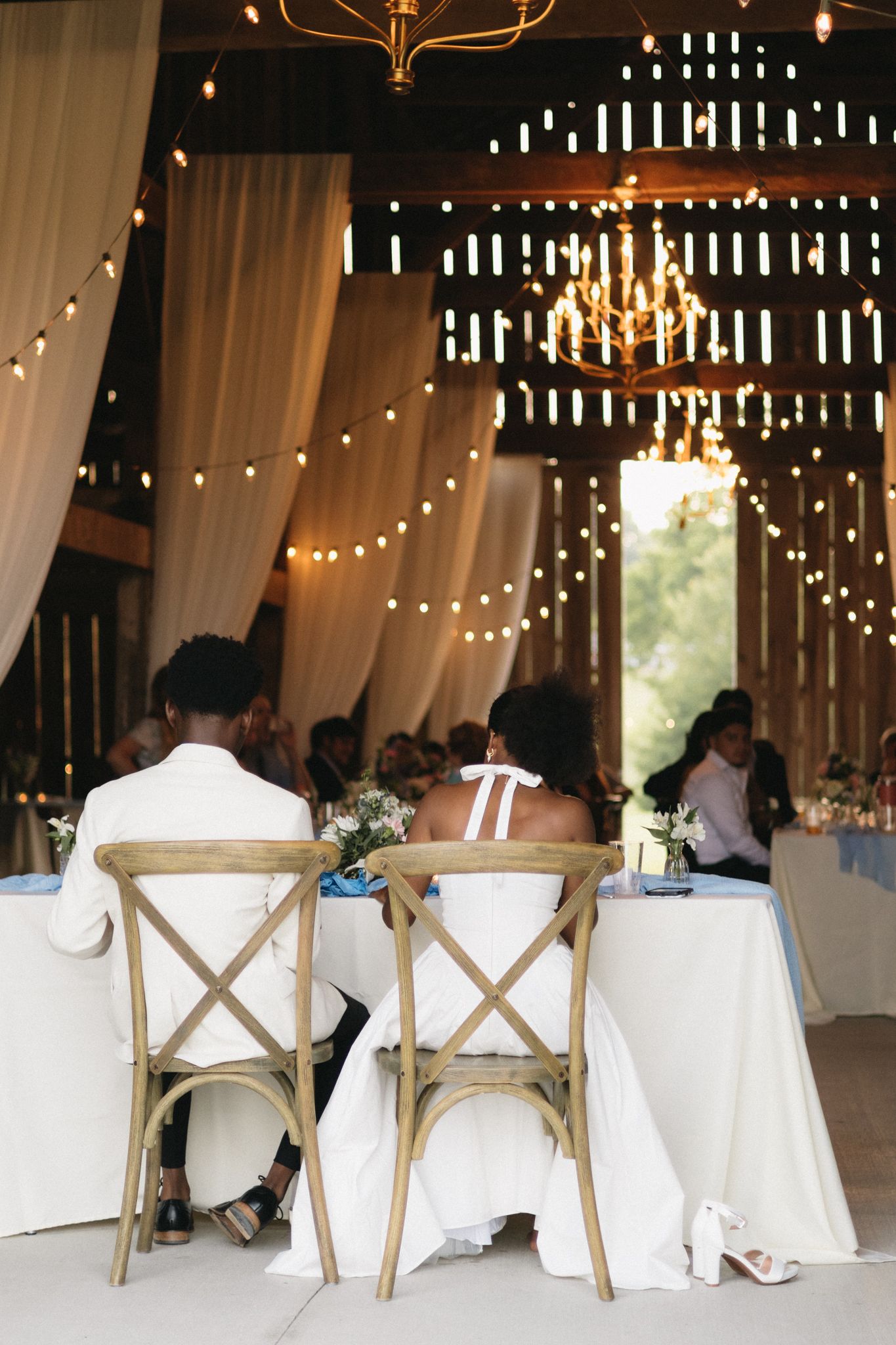 Rustic barn wedding reception with string lights, white drapes and cross-back chairs at a head table.