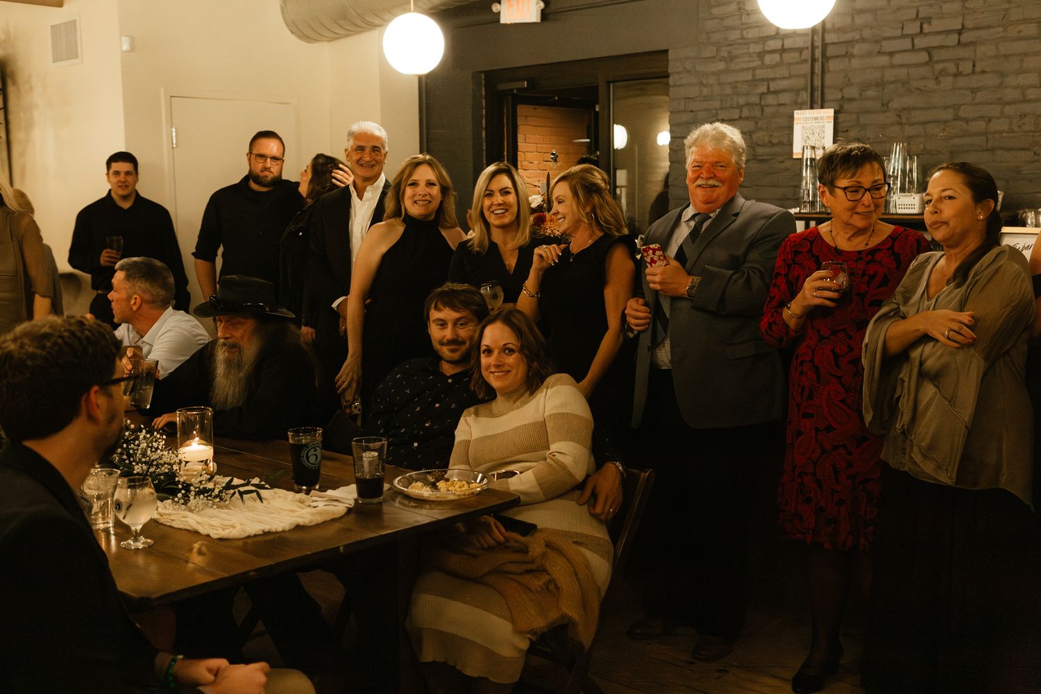 A warmly lit restaurant gathering of people standing and sitting around a table in the evening.