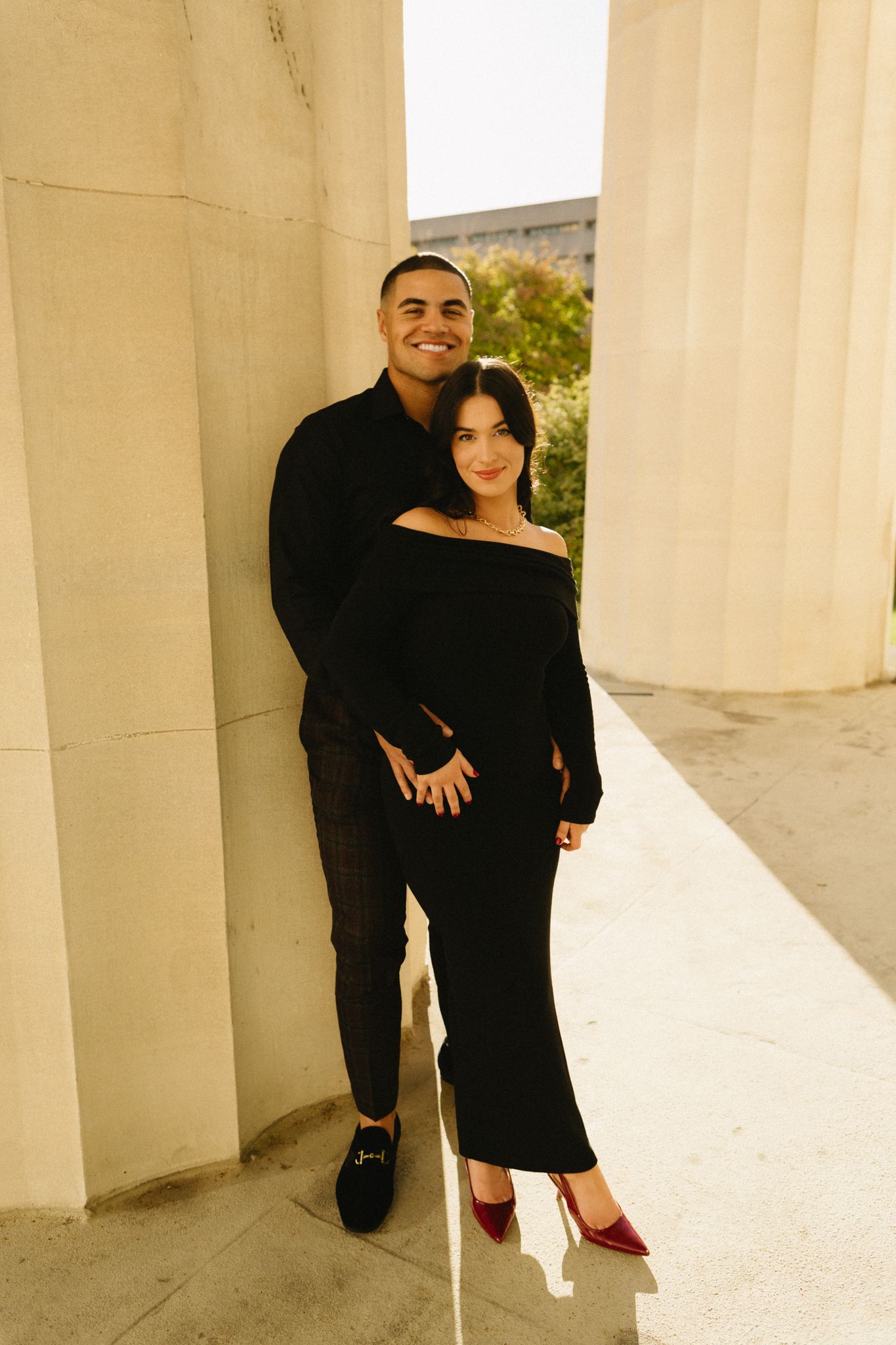 A stylish couple in matching black outfits poses together between classical columns outdoors.